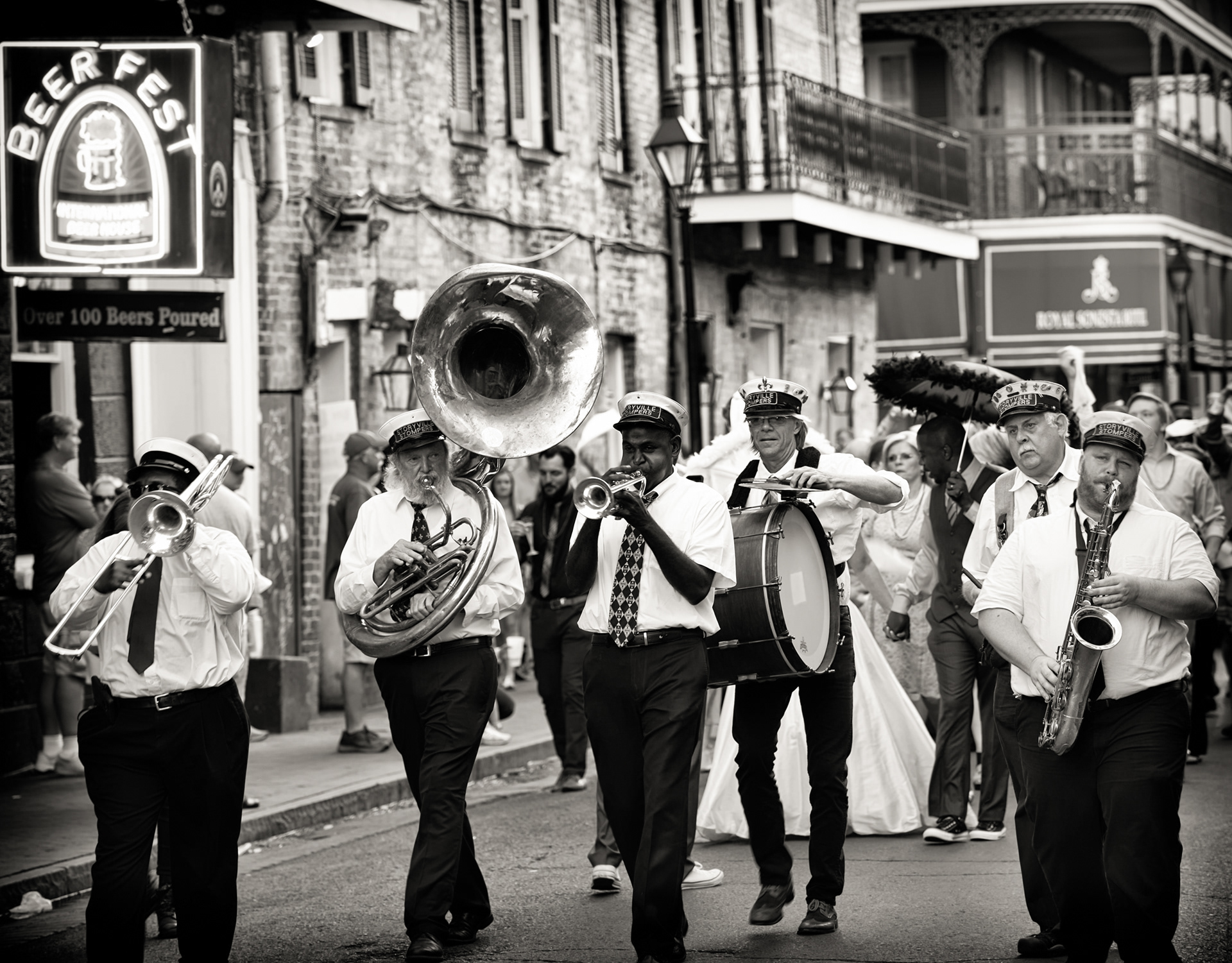 Their hats say Storyville Stompers — the band's actual name, thirty years and six thousand gigs in — leading a wedding second line down a French Quarter street: tuba, trombone, trumpet, snare, sax, the bride trailing behind in white under an umbrella that's tradition, not rain gear. New Orleans brass bands trace to emancipation-era parade and funeral societies, and jazz grew straight out of that sound. A band leading a bride and groom past a Beer Fest sign isn't a novelty for tourists — it's the same tradition, just pointed at a wedding instead of a funeral.