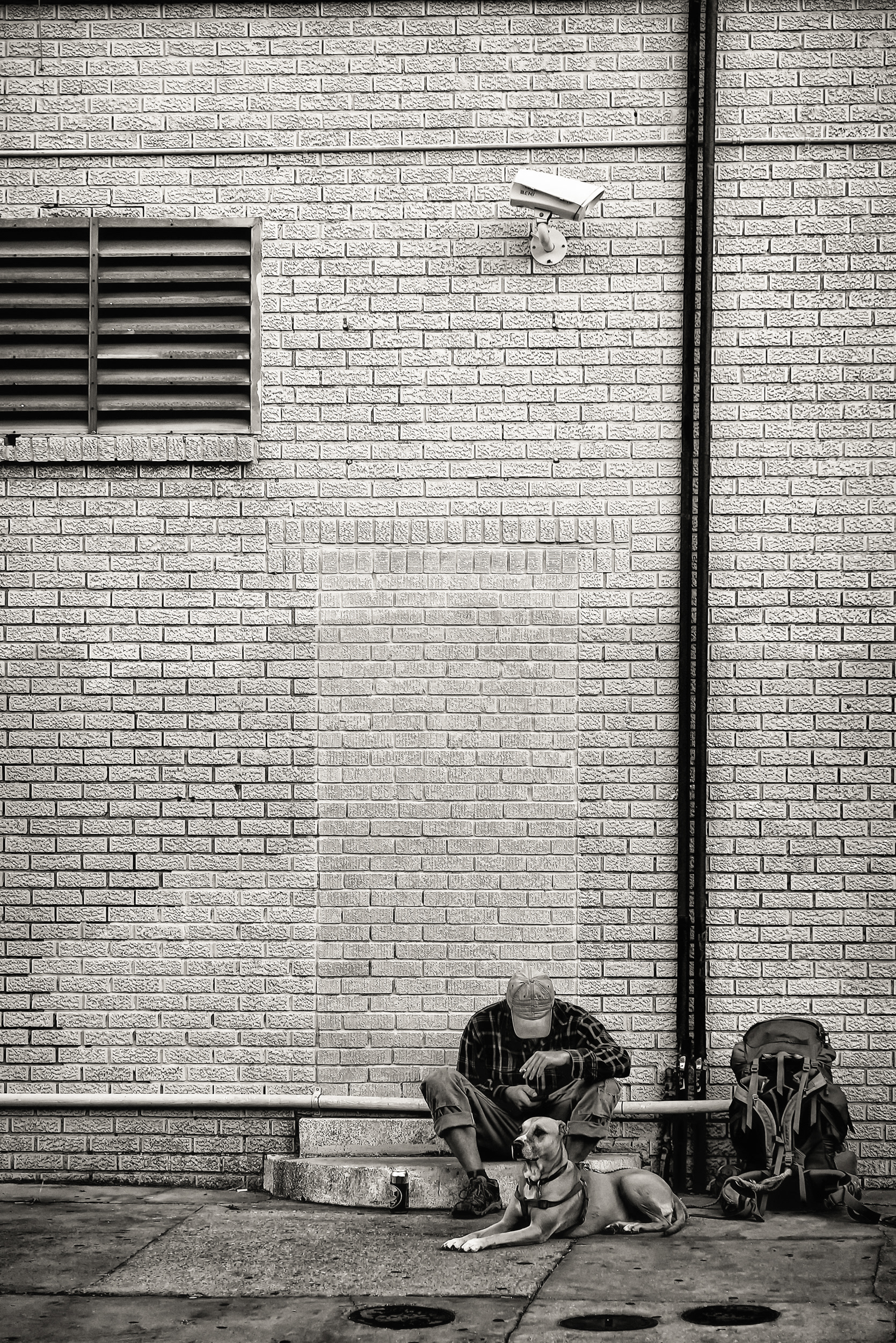 A man and his dog on a stoop, backpack beside them, a security camera watching from above. I don't know their story — just the irony of the camera