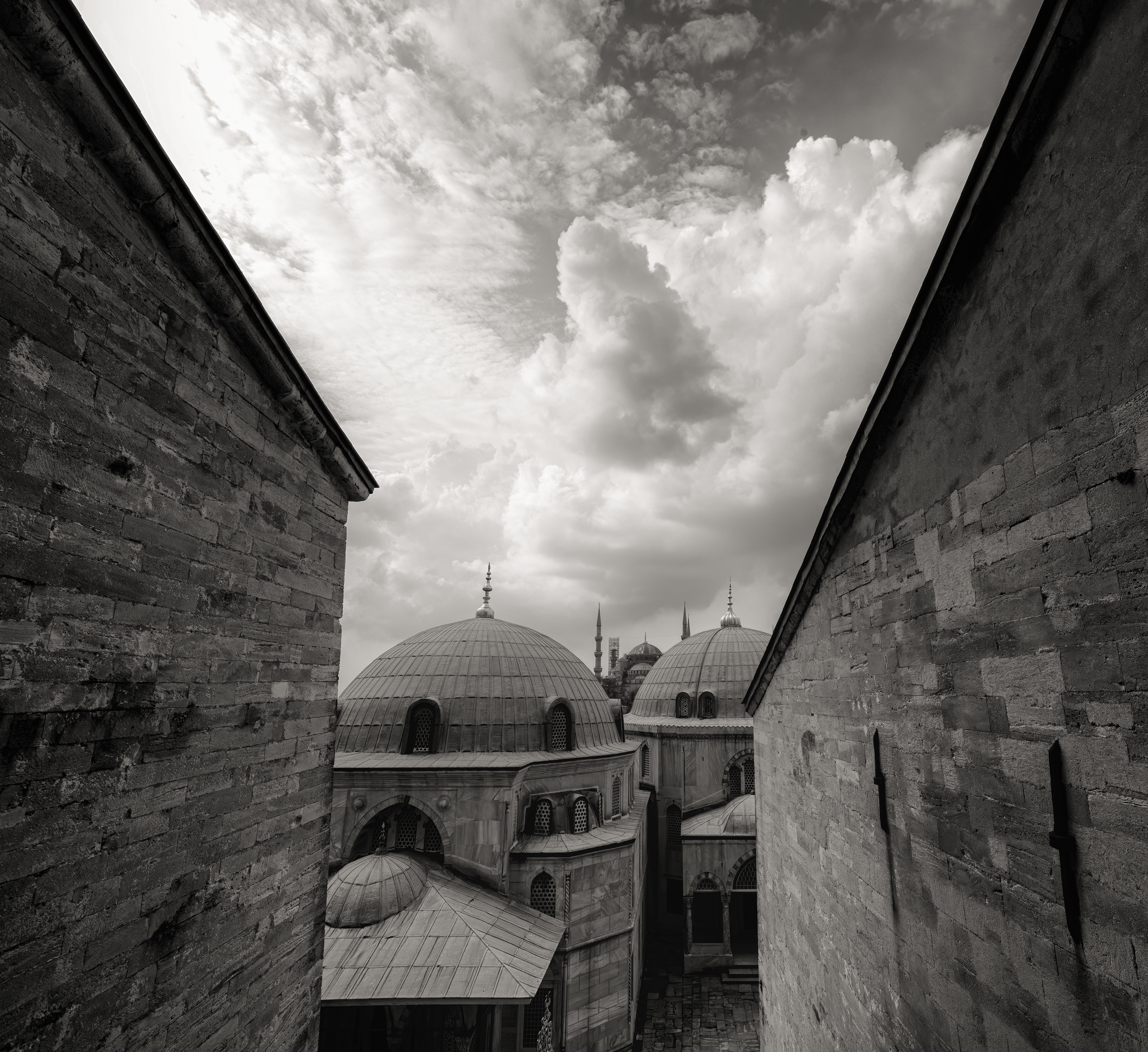 Domes, storm clouds moving through, black and white. Hagia Sophia's cascading domes and semi-domes — one central dome stepping down into smaller ones, buttressed outward in tiers — became the template Ottoman architects reused for centuries afterward, the Blue Mosque included. Different faiths, same skyline logic, borrowed wholesale.