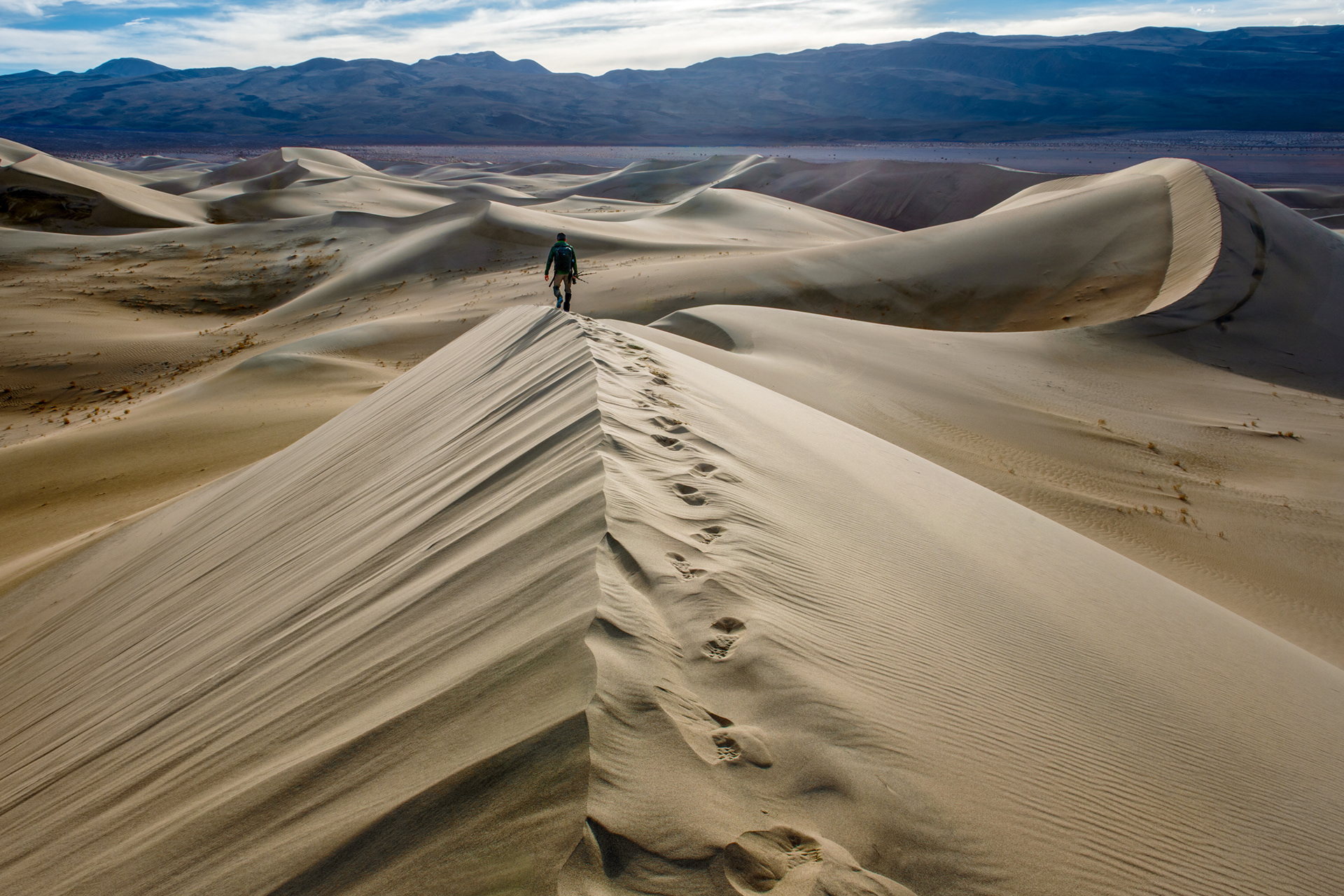 Mesquite Flat, Death Valley, late afternoon, the ridgelines throwing the kind of shadow only a low sun gives you. Despite the name of the park, dunes like this are rare here — it takes loose sand, wind to move it, and mountains on both sides to keep it from just blowing away, and that combination barely exists in Death Valley at all. One figure walking the ridge, one line of footprints the wind will have opinions about by morning.