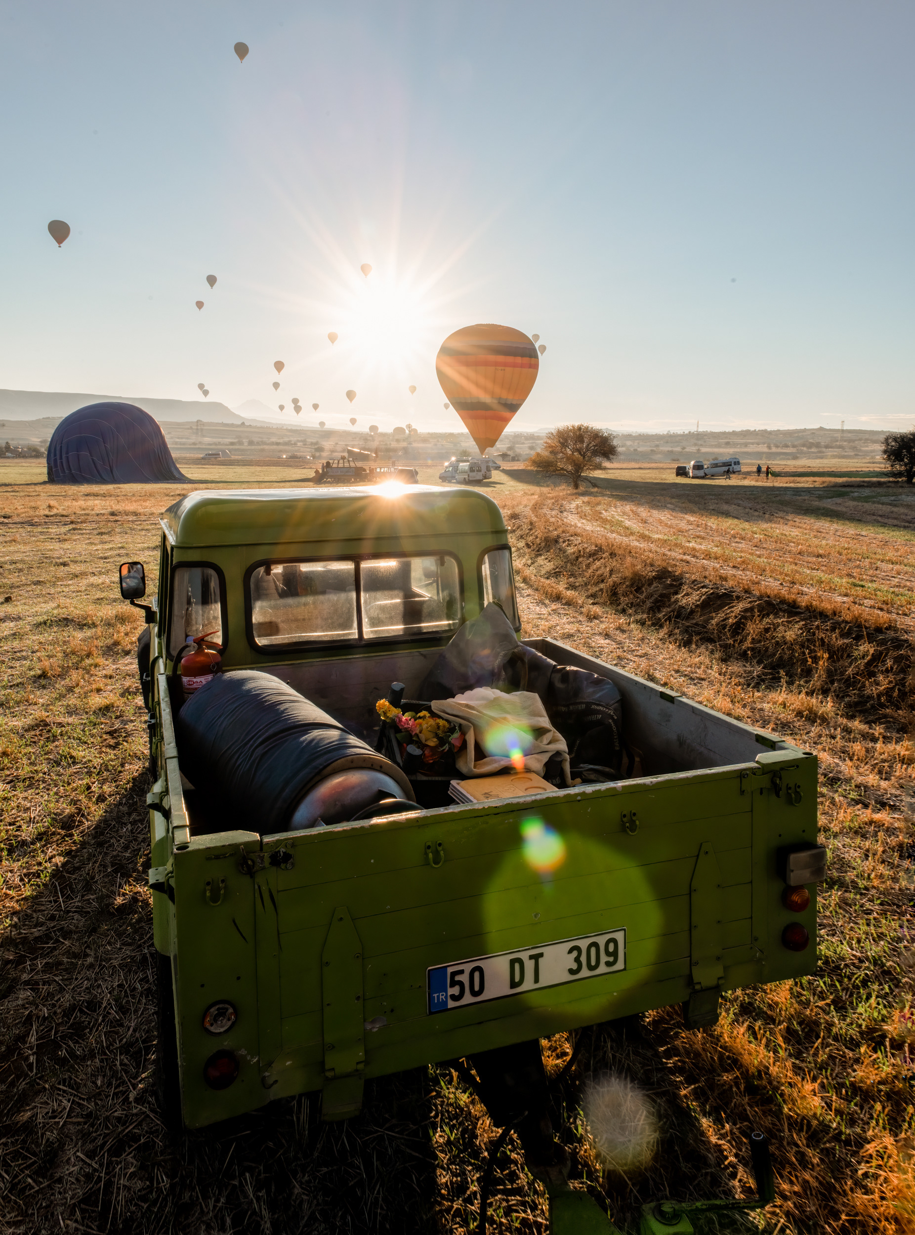 A hot air balloon has exactly one control: up or down, by how hard the burner fires. Everything sideways is the wind's decision, not the pilot's, which makes the chase crew's whole job a guessing game — no flight plan, just a radio and whoever's driving learning to read the same sky the pilot's reading, trying to reach the landing spot before the balloon does. First-timers traditionally get a certificate afterward, named for the Montgolfier brothers, who managed the first untethered flight in 1783 without so much as a truck waiting on the other end. This one's already landed light on ceremony: a bouquet left in the truck bed, one more balloon still working its way down behind it, the morning quietly wrapping up.