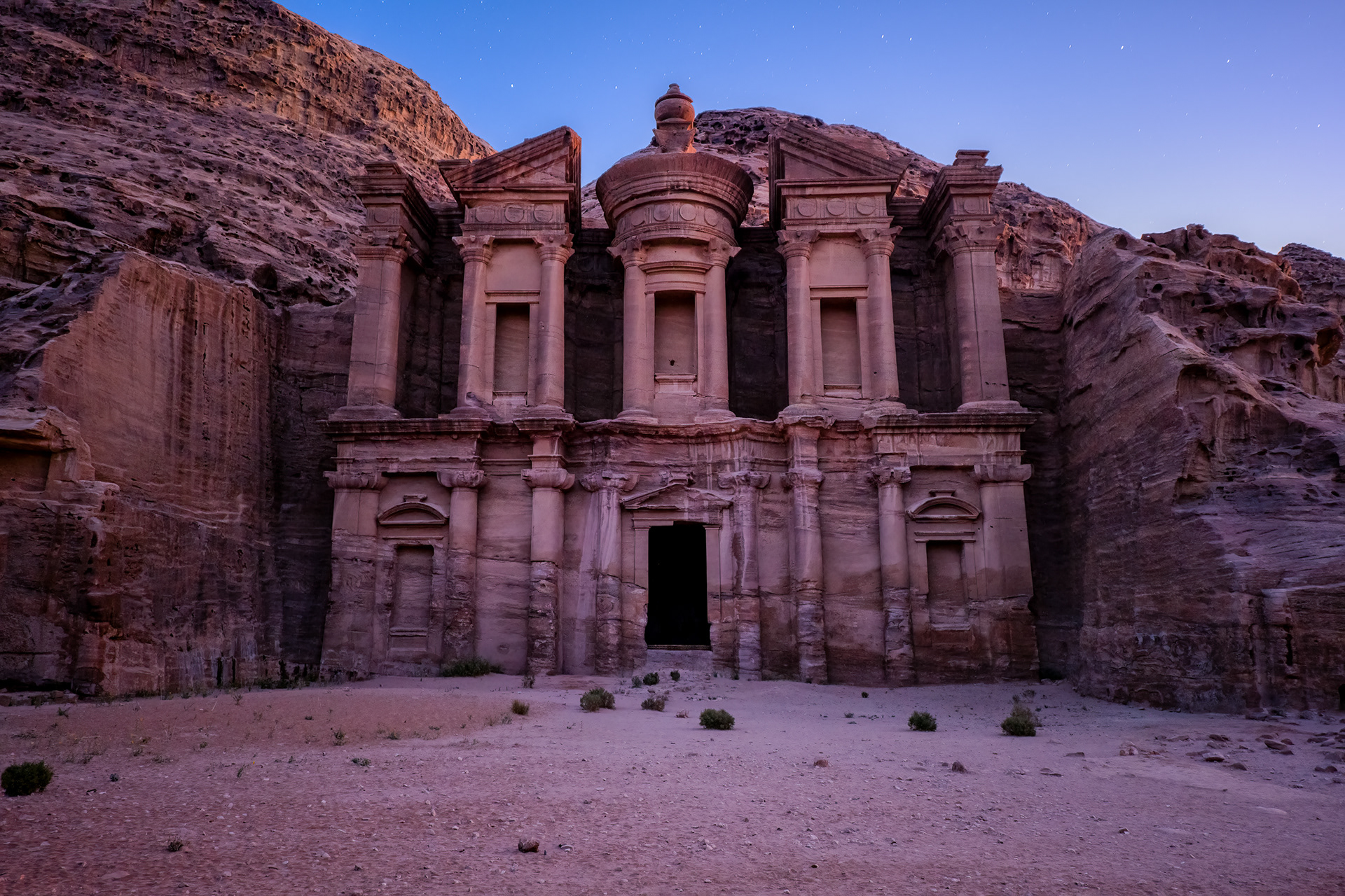 The Monastery. Carved in the first century, a hall for a dead king's feast. Later the monks came and cut crosses into the stone, and it took a new name — Ad Deir. I went up in the dark, before the sun, eight hundred steps of it. Women sold silk scarves out of the shadows. At the top, the façade stood in the last blue light.