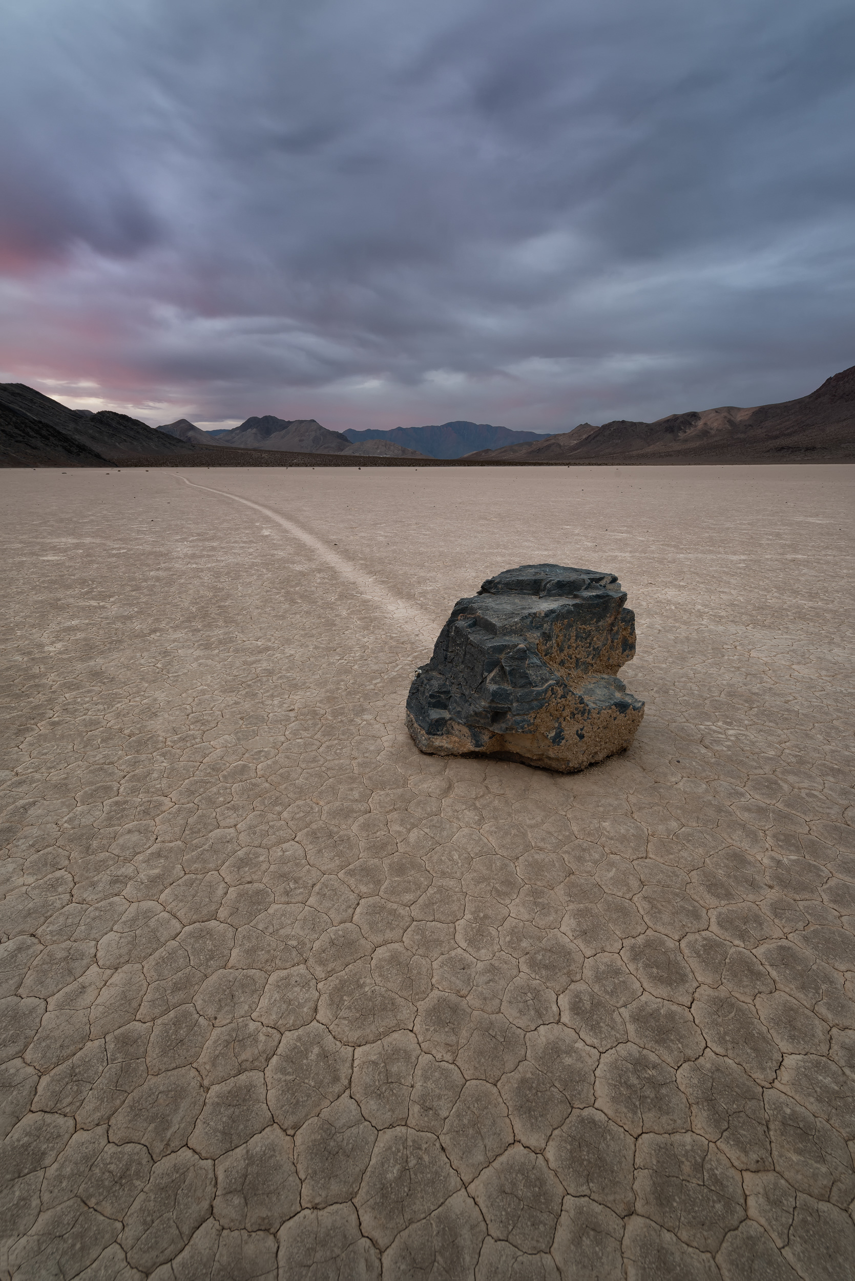 Racetrack Playa, dawn, a 4-second exposure to pull enough light out of the sky. The rocks here move on their own — trails scratched into the mud stretch up to 1,500 feet — and nobody actually knew why until 2014. Turns out it takes a thin sheet of ice from an overnight rain, morning sun to melt it just enough to float, and wind to do the pushing. Take away any one of the three and the rock just sits there being a rock, the way it does everywhere else on Earth.