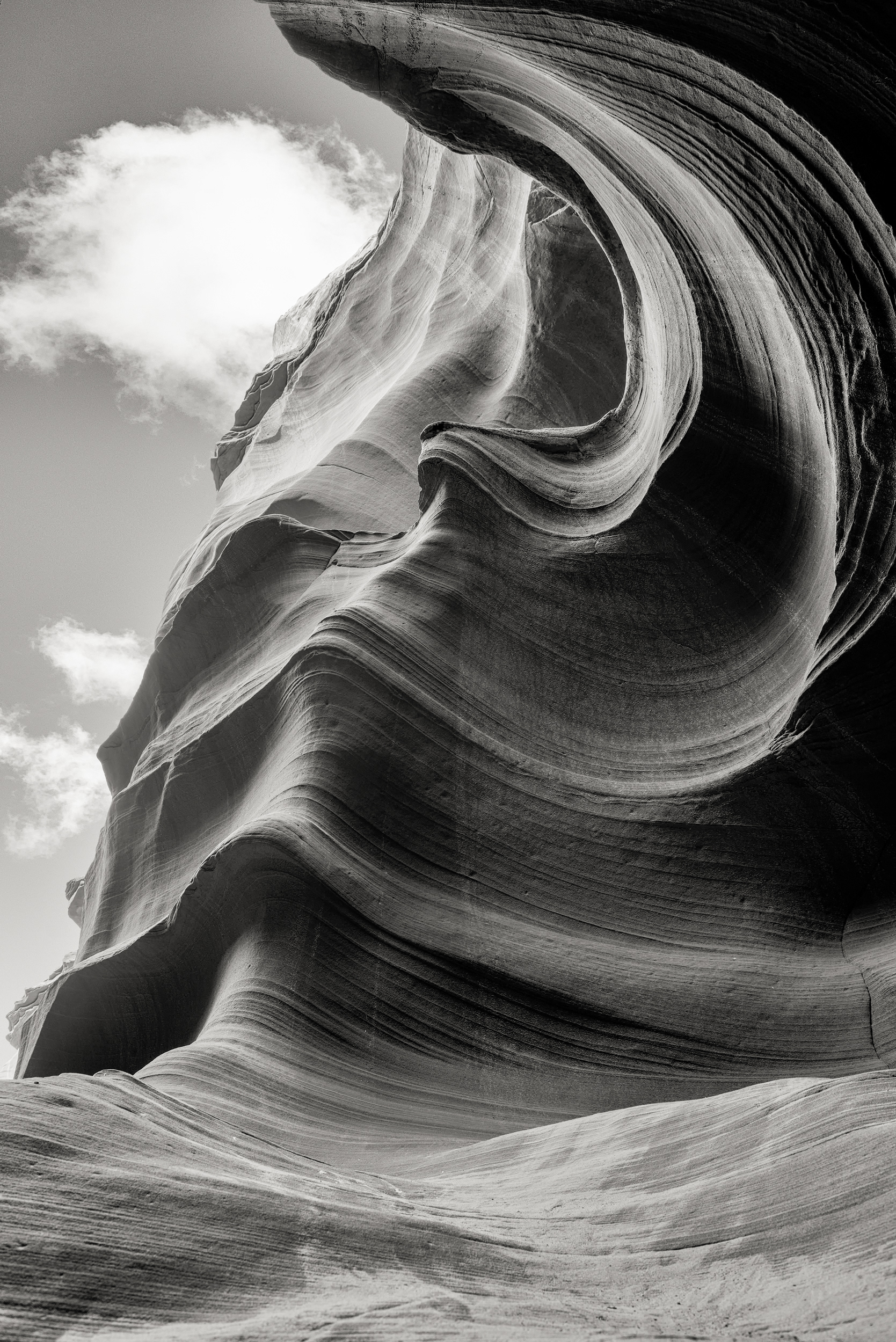 Slot canyon, a single cloud caught in the narrow ribbon of sky overhead, black and white — and what's genuinely strange, standing at the bottom of one of these things, is the way the sky stops functioning as background and becomes instead this very specific, very thin, almost architectural event happening directly above you, so far up and so hemmed in on both sides that one drifting cloud, for however many seconds it's actually in frame, becomes more or less the only thing happening anywhere — a kind of forced, involuntary attention you don't really get anywhere else, the visual equivalent, maybe, of staring at a single candle flame in an otherwise dark room, i.e., not because the cloud itself is remarkable but because two hundred vertical feet of sandstone has quietly removed every other possible object of your attention.