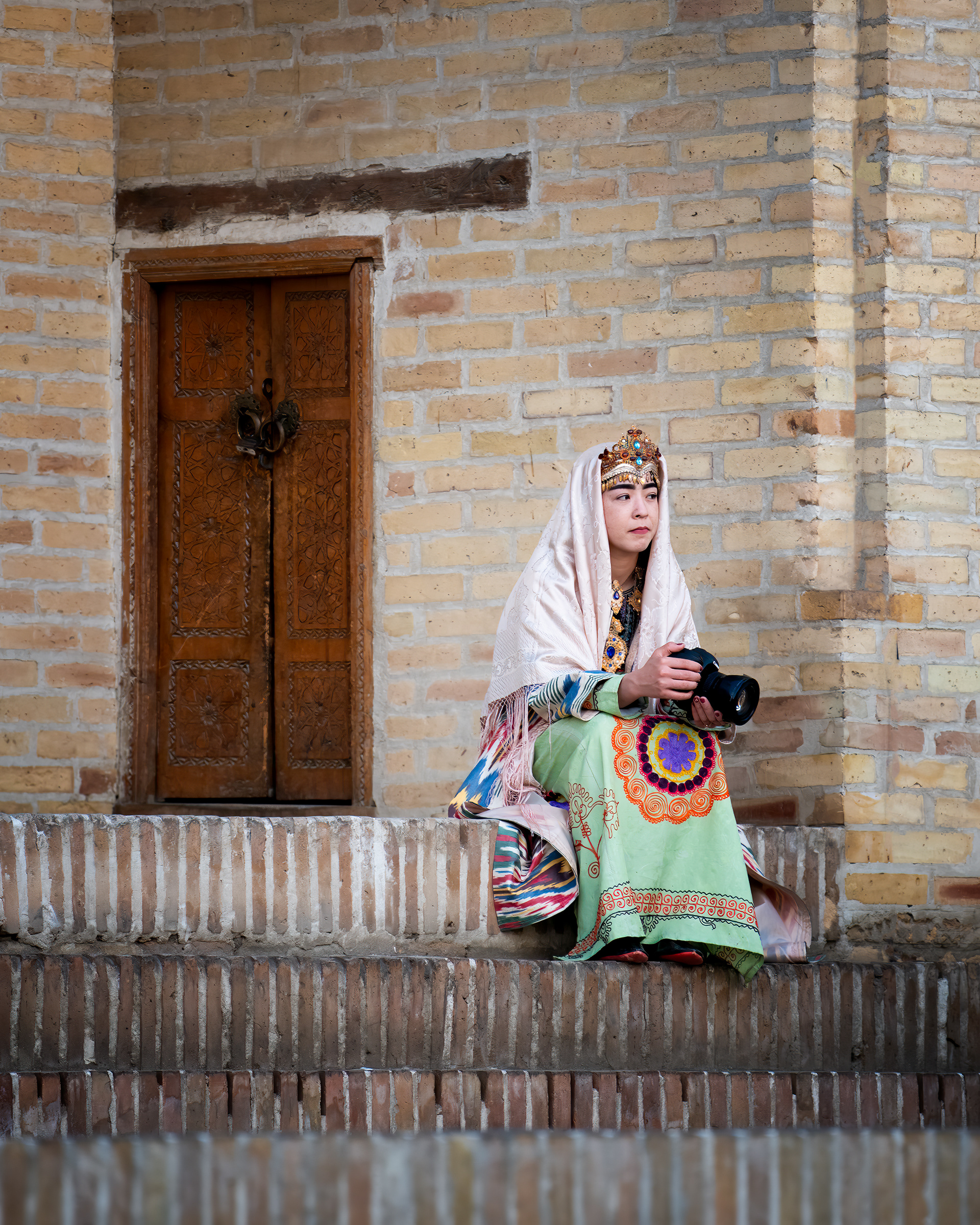 She's dressed like she stepped out of a miniature painting — the crown, the ikat robe, the carved door behind her doing its part — except for the camera in her hands, which is newer and probably better than mine. I don't know if that's irony or just what tourism looks like now: the past gets put on as a costume, and everyone still brings their own gear.