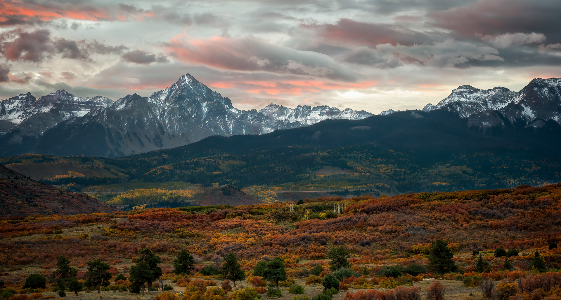 The Sneffels Range at dusk. Mount Sneffels itself, the 14,150-foot peak anchoring this skyline, is named after Snæfellsjökull, the glacier-capped Icelandic volcano that Jules Verne used as the entrance to the underworld in Journey to the Center of the Earth — an oddly specific reference for nineteenth-century surveyors to reach for out here, but that's the naming. This particular vantage, from along Highway 62 at the Dallas Divide, is reportedly one of the most photographed mountain views in the state, and as of 2023 it's also the peak printed on every Colorado driver's license — which means more people are now carrying around a version of this exact mountain in their wallet than have ever actually stood here and seen it.