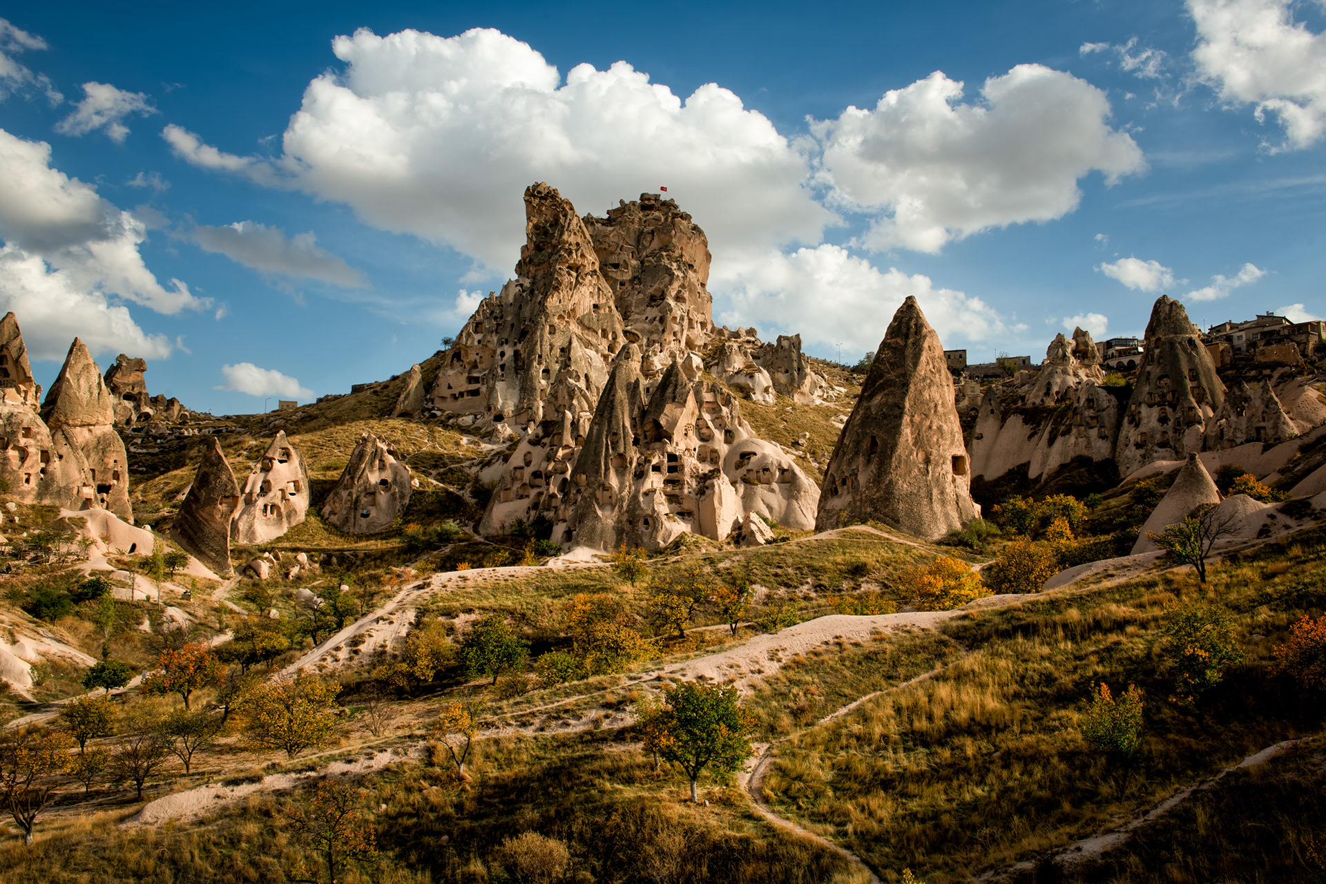 Fairy chimneys — peri bacaları, in Turkish, and that's the literal term geologists use for them too — each one a soft cone of compressed volcanic ash laid down by eruptions millions of years ago, capped by a disc of harder rock that erodes slower than everything beneath it. That mismatch is the whole shape: the cap shields a shrinking pedestal the way an umbrella shields the ground under it, right up until erosion undercuts the pedestal too far and the cap finally tips off — at which point the chimney underneath is just as exposed as its bare, capless neighbors and starts wearing away for good. The ones still standing this tall simply held onto their caps longer. The flag on top isn't geological. That's just Turkey, claiming the highest available point, the way it does on ridgelines and minarets across the whole country.