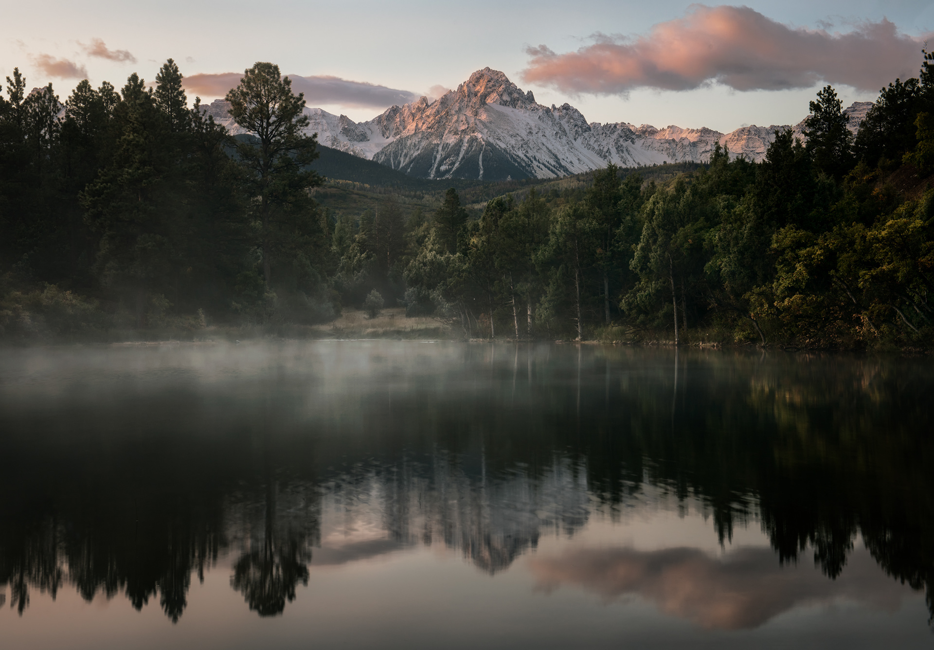 Foggy dawn, lake reflection. Both things in this frame come from the same overnight condition: clear skies and dead-calm air let the valley floor lose heat fast after dark, cold air then drains downhill and pools right where the lake sits, cooling below its dew point and condensing into exactly this kind of low-lying fog. That same stillness is also what makes the reflection possible — glassy water needs almost no wind at all, and a valley cold enough to fog over is, by the same physics, usually calm enough not to disturb the surface. By midmorning, once the sun's warmed the air past that threshold, both effects are gone within the same half hour.
