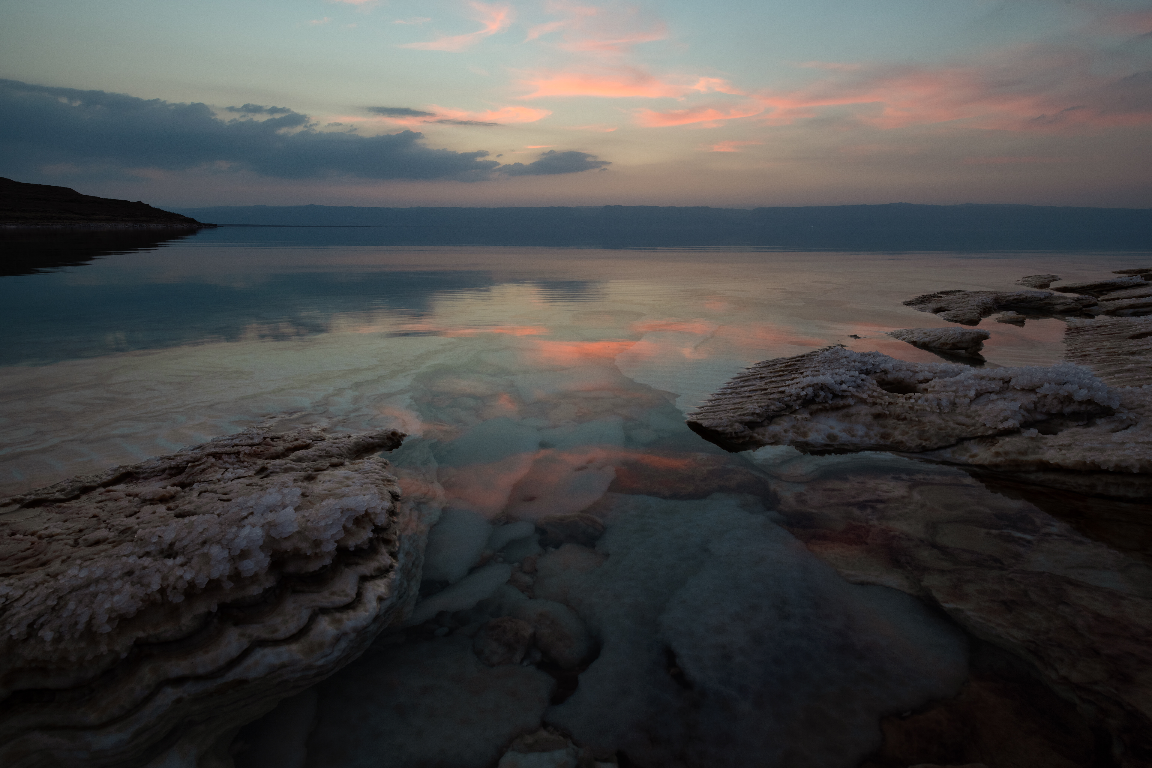 Dead Sea, dusk. Salt crusts the rocks like frost that never melts. Across the water, just visible in the haze, sits the West Bank. The sky went pink, and the sea held still enough to show it twice