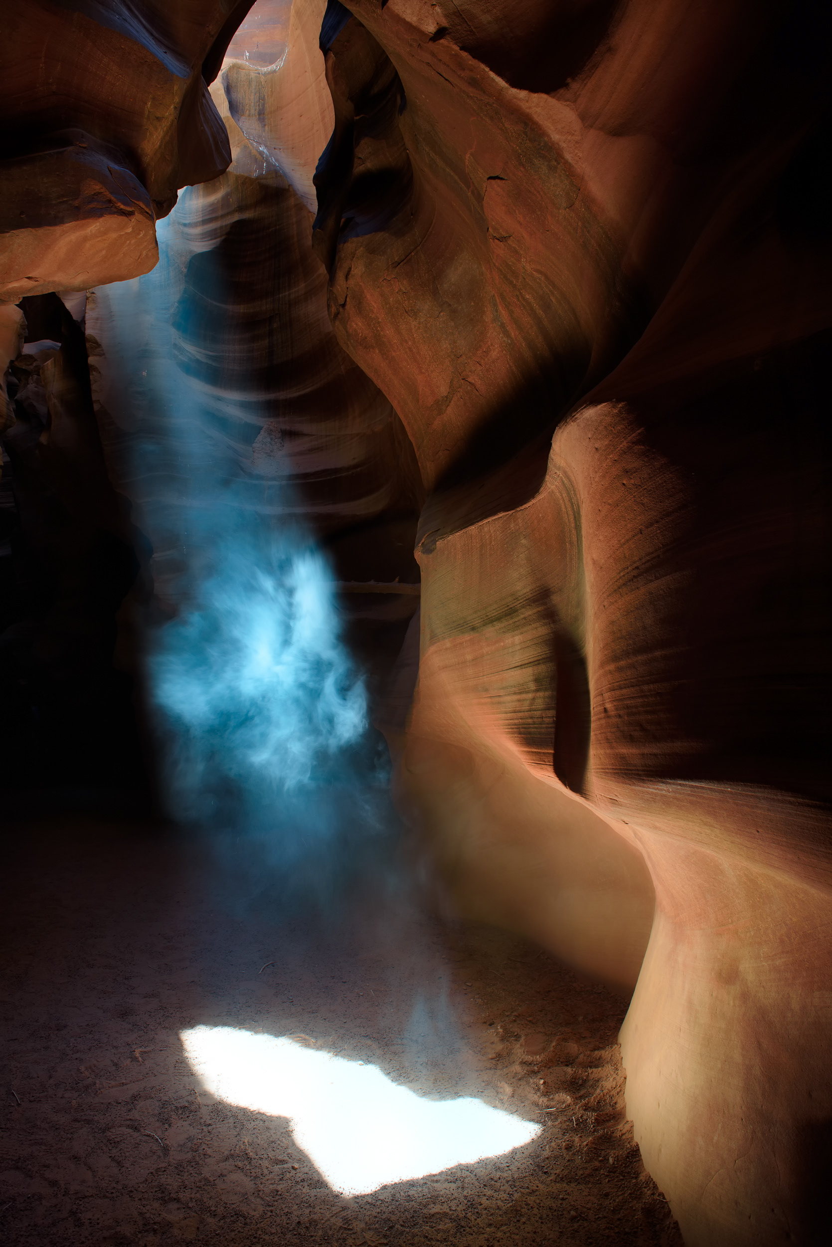 Slot canyon, a shaft of light reaching the floor. A beam like this only becomes visible if there's something in the air for it to hit — dust, mostly, kicked up naturally or, more often, tossed by a guide with a handful of sand at the right moment, since light scattering off airborne particles (the same effect that makes headlights visible in fog) is the only reason you see a shaft instead of just a bright patch on the ground. It also only reaches the floor when the sun sits nearly straight overhead, which narrows the window to a couple of hours around midday, and only for a few months out of the year when the sun's angle cooperates with how narrow this particular slot runs.