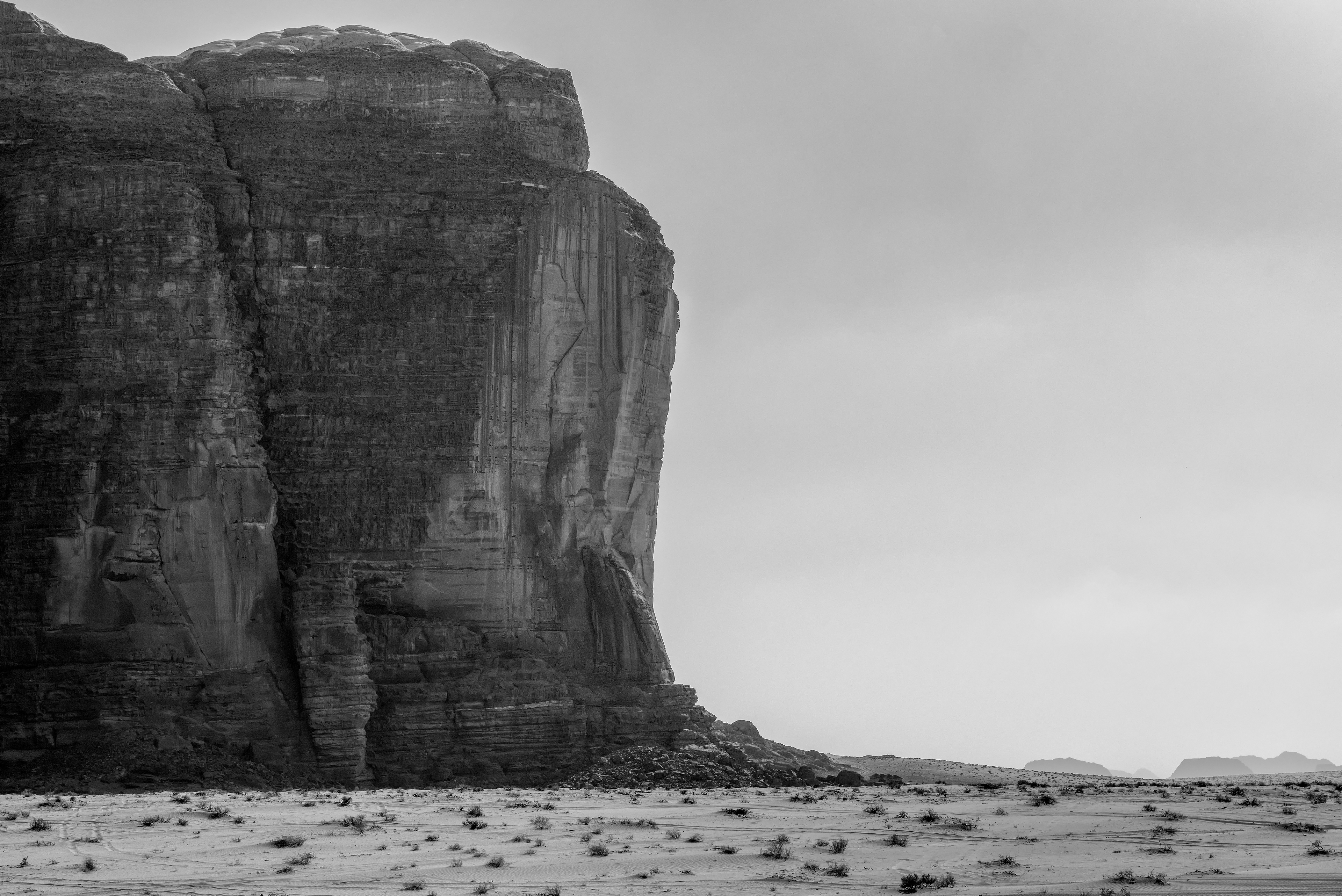 Sandstone monolith, black and white — Wadi Rum. A weathered cliff face streaked dark with desert varnish against an empty, hazy sky.