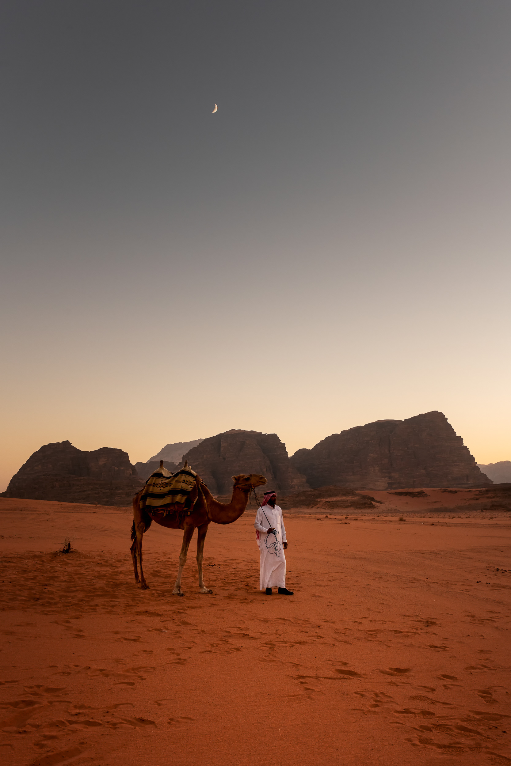 Man, camel, and crescent moon — Wadi Rum. Called the "ship of the desert," the camel made life here possible for thousands of years, when nothing else could cross this sea of sand. Man and camel stand together in the day's last warmth, a crescent moon rising above them.