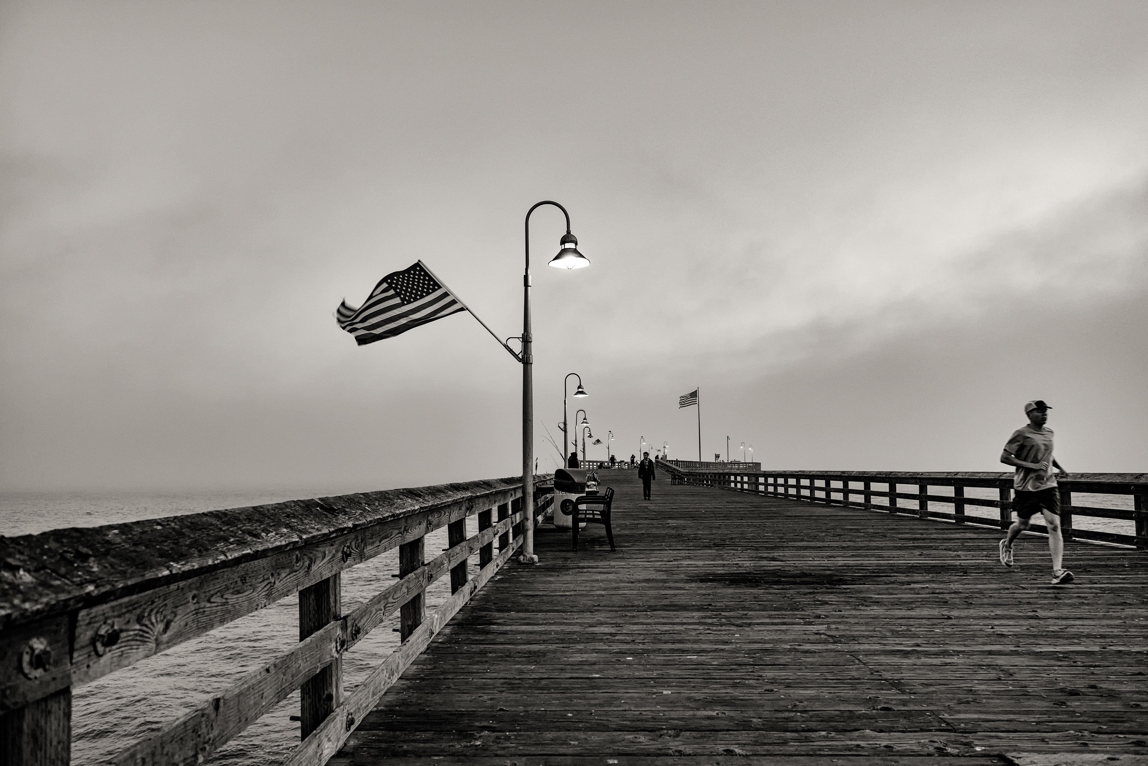 Ventura Pier, evening fog, black and white. Built in 1872, first as a commercial wharf for shipping lumber, produce, and crude oil out before roads made that unnecessary. Once California's longest wooden pier, just under 2,000 feet — until a December 1995 storm sheared off 420 feet of it in a single night, on top of a ship fire, a steamer collision, and half a dozen other storms across the century before that. What's left still stands, now used for fishing, walking, and, here, a jog through the fog