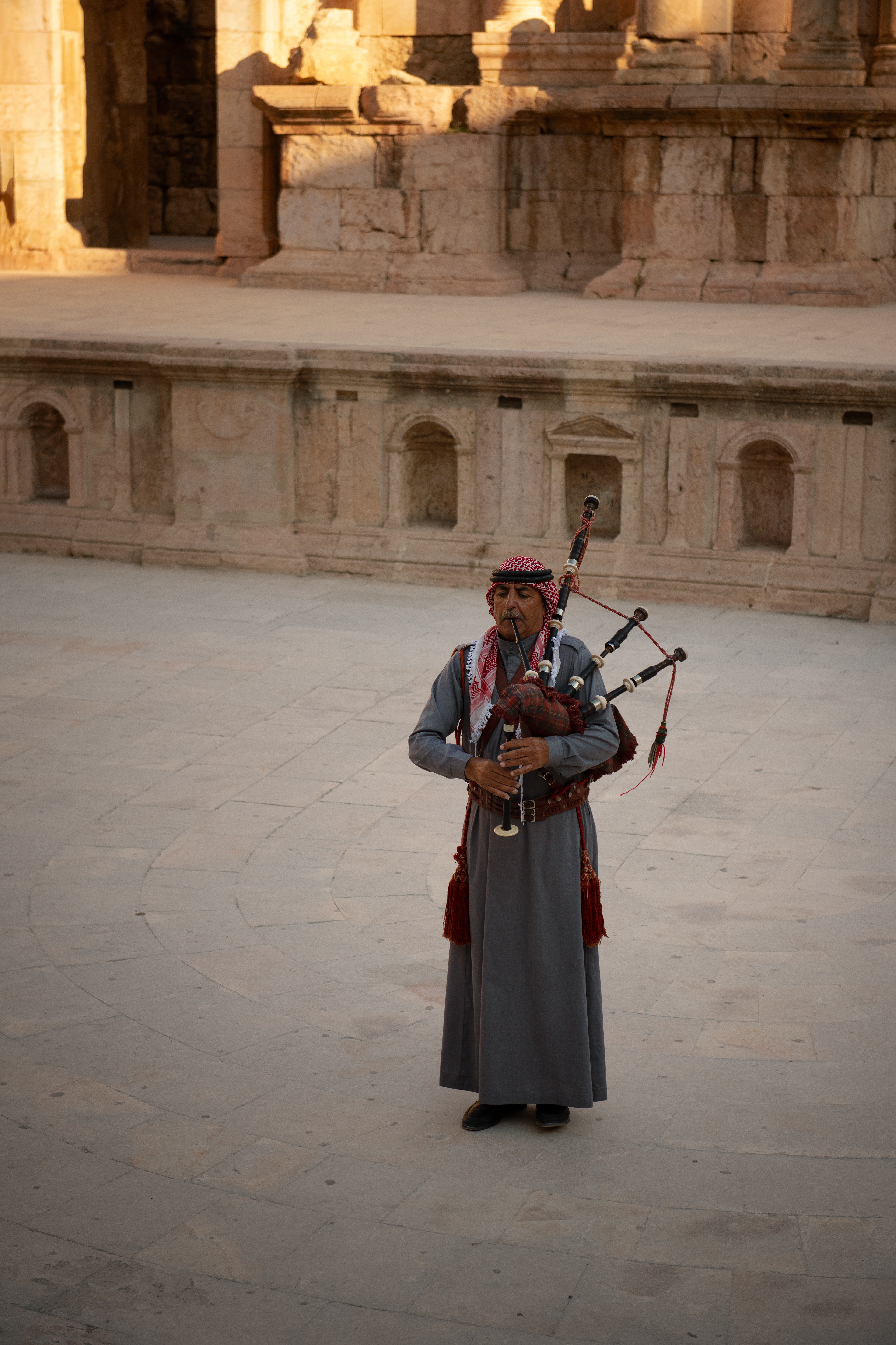 Jerash. A piper in keffiyeh, standing alone on the stage of a Roman theater. The British brought bagpipes here in the 1920s while training Transjordan's army; the Mandate ended, the pipes stayed, and they've been Jordan's own ever since.