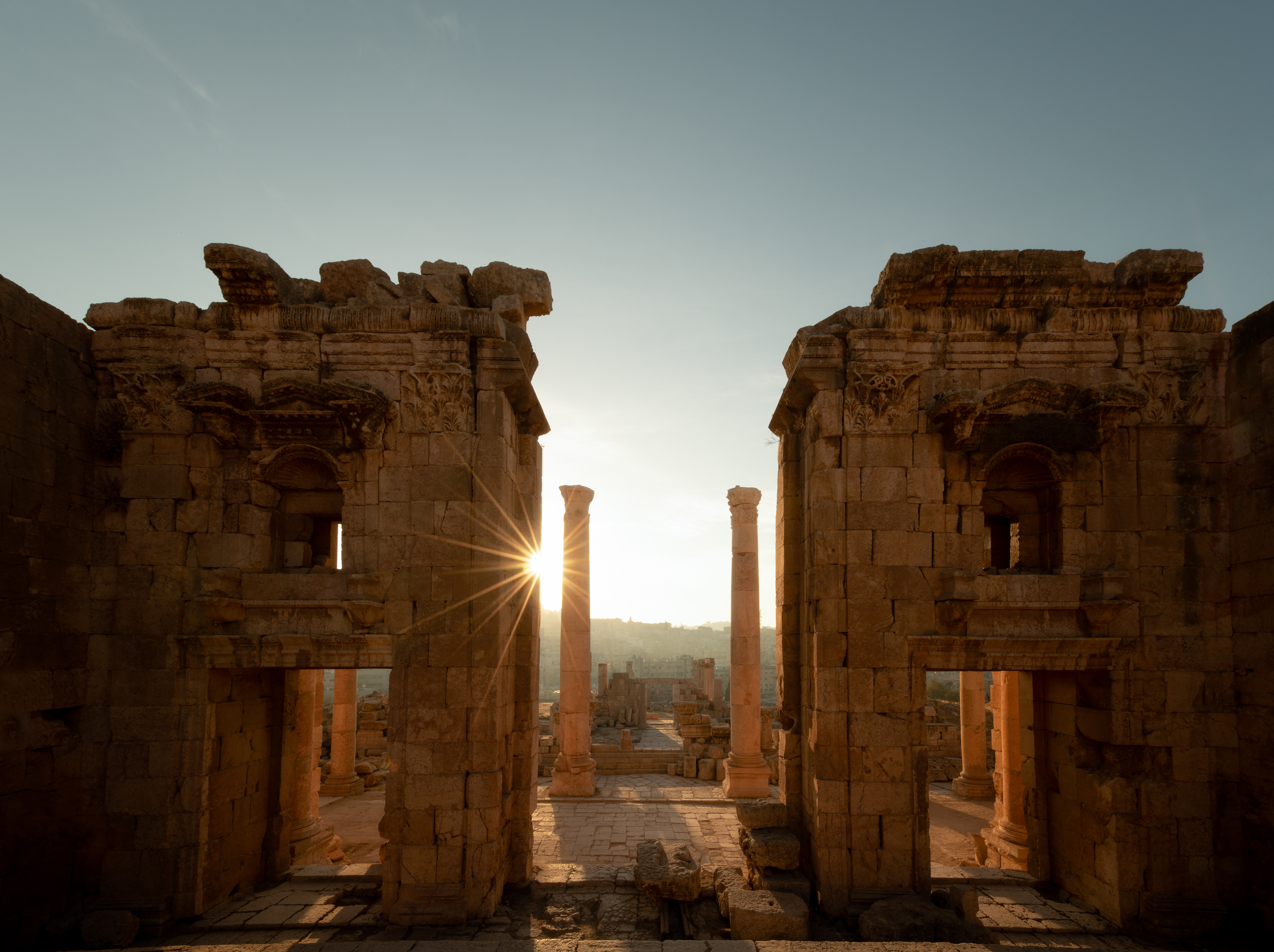 Jerash, the Temple of Artemis — built for the city's patron goddess, and never finished. The columns aren't rigid: stacked stone drums pinned on an iron dowel, built to sway in an earthquake rather than crack. The sun found the gap between them, the modern city waiting just beyond.