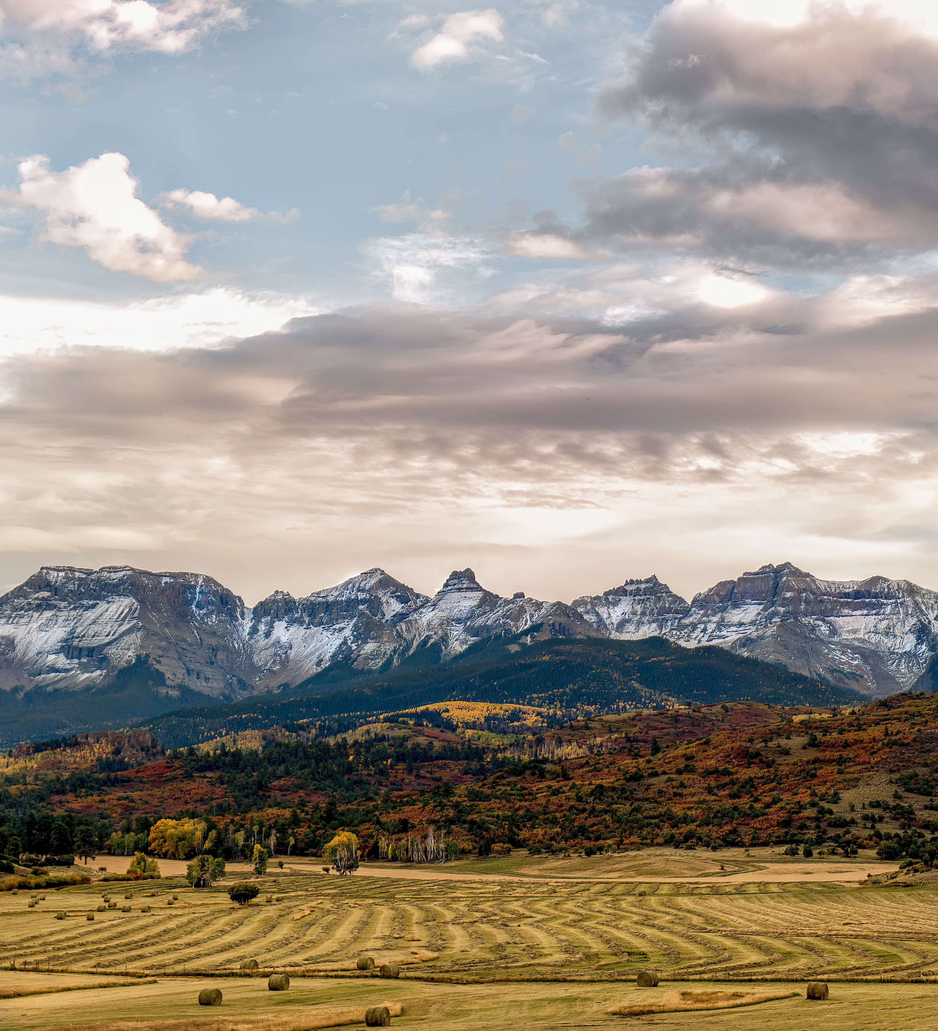 Hay rows, cut and raked into windrows below the peaks. At this elevation the growing season runs short enough that most of these meadows get exactly one cutting a year, timed to a narrow window in mid-to-late summer between the last frost and the first one. The rows themselves are windrows, not the original cut pattern — a separate pass with a rake gathers the hay into lines like this specifically so it dries evenly before the baler comes through, which is also why the spacing looks so deliberate: it's set by the width of the equipment, not by anyone drawing lines in a field for the view