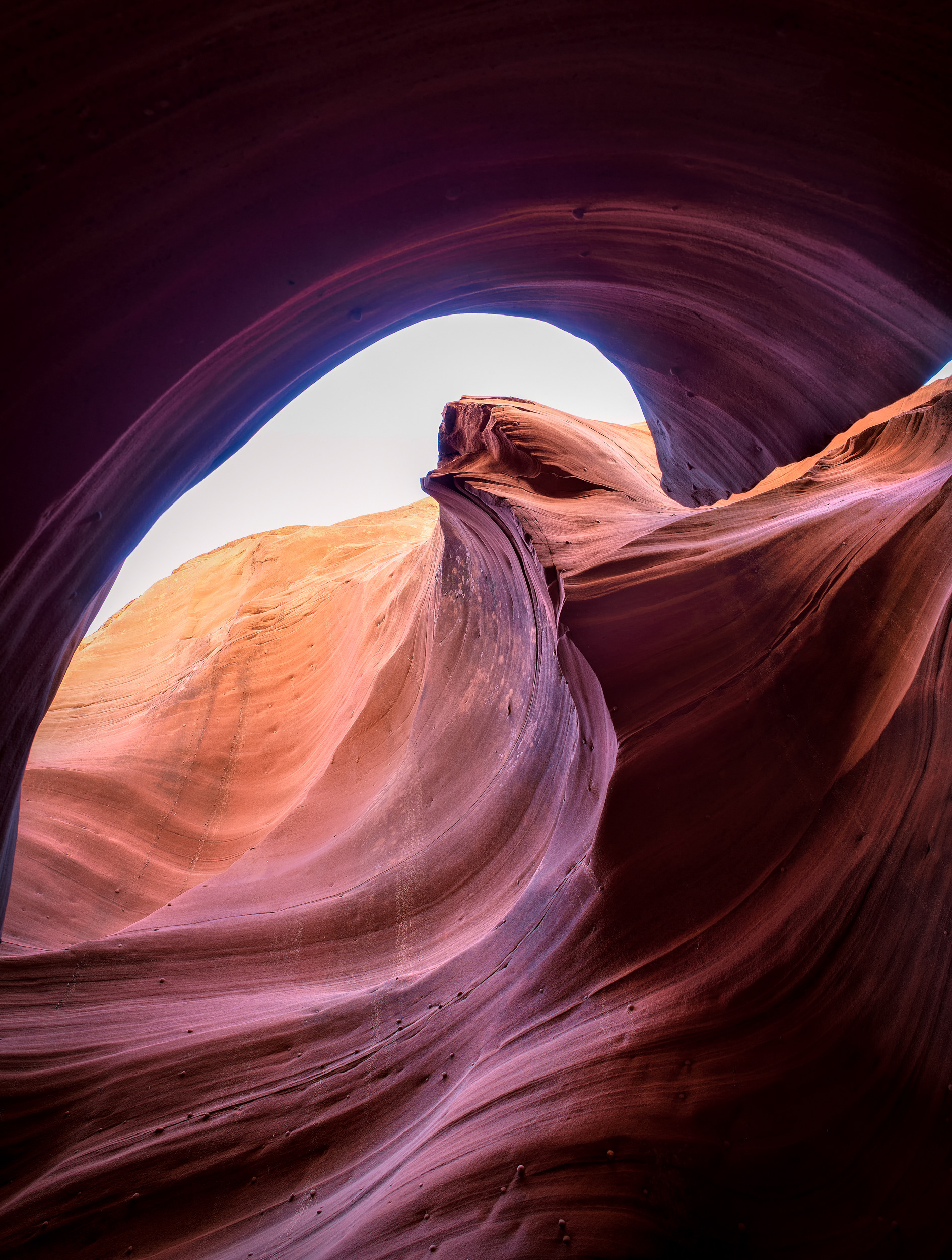 Slot canyon, an arch framing a portal further down the passage. Water carved this, not wind — flash floods through passages like this one carry enough sand to sandblast the walls smooth with every real rain, which is also why the shapes keep changing over time: a single storm can cut a curve that didn't exist the week before. Most of Arizona's classic slot canyons, this one included, sit on Navajo Nation land and require a Navajo guide to enter — partly cultural, partly because these passages take flash floods seriously; a nearby slot canyon of the same type killed eleven visitors in a single 1997 flood, and every access system since has been built around not repeating that.