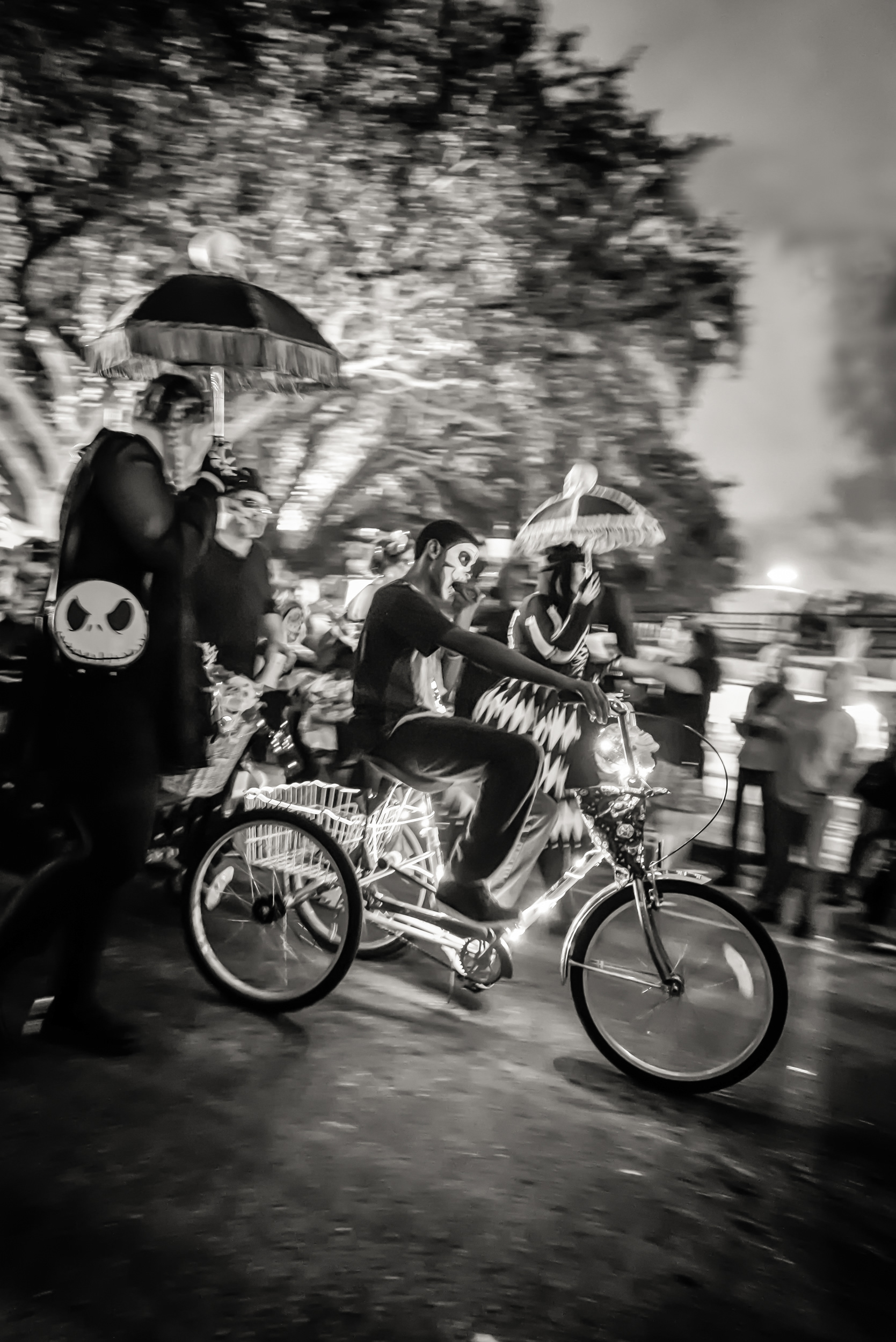 A man pedals a decorated tricycle through the crowd, face painted into a skull, and he's not even the densest part of the frame — there's a parasol behind him, another parasol-and-skeleton-mask combo to his right, a zebra-striped harlequin dress somewhere in the mix, which is to say the actual subject here might just be how much costume a single frame can hold before it stops registering as "a lot" and starts registering as, simply, the ambient condition of the evening (there's a term for this in crowd photography, probably, though I don't know it)