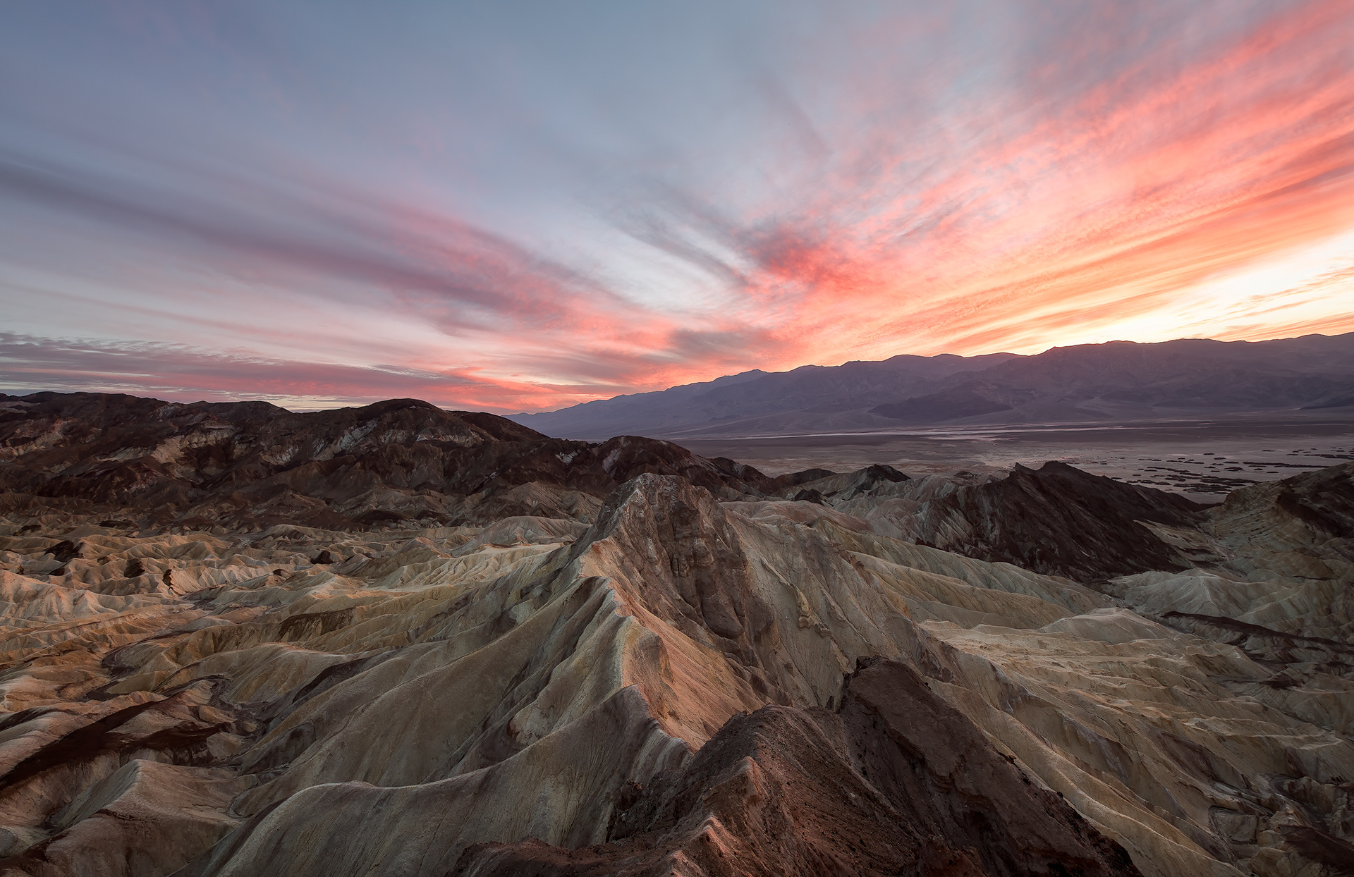 Zabriskie Point, sunset, Death Valley — shot from a ridge above Golden Canyon, near Red Cathedral, well off the paved overlook everyone else uses. These badlands are the floor of a lake that started filling in roughly nine million years ago: saline mud, mountain gravel, layer after layer of volcanic ash, all left high and dry once the climate turned and the lake just stopped existing. Manly Beacon stands taller than everything around it not because the rock's any tougher, but because a hard cap of 3-to-5-million-year-old lava happened to land on it and slow the erosion down. Named for Christian Zabriskie, an executive at the borax company whose twenty-mule teams once hauled ore out of this same valley