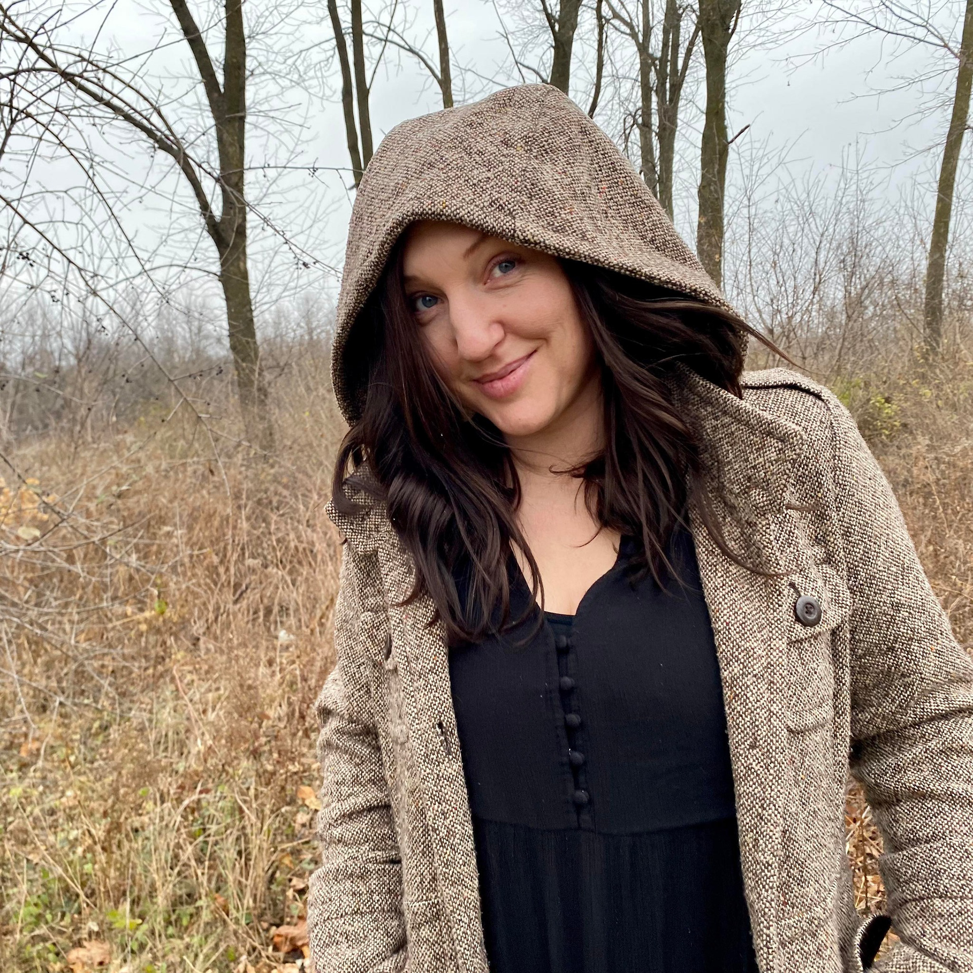 Desirae Hill smiling looking at camera, wearing a long brown coat and black dress standing in the woods