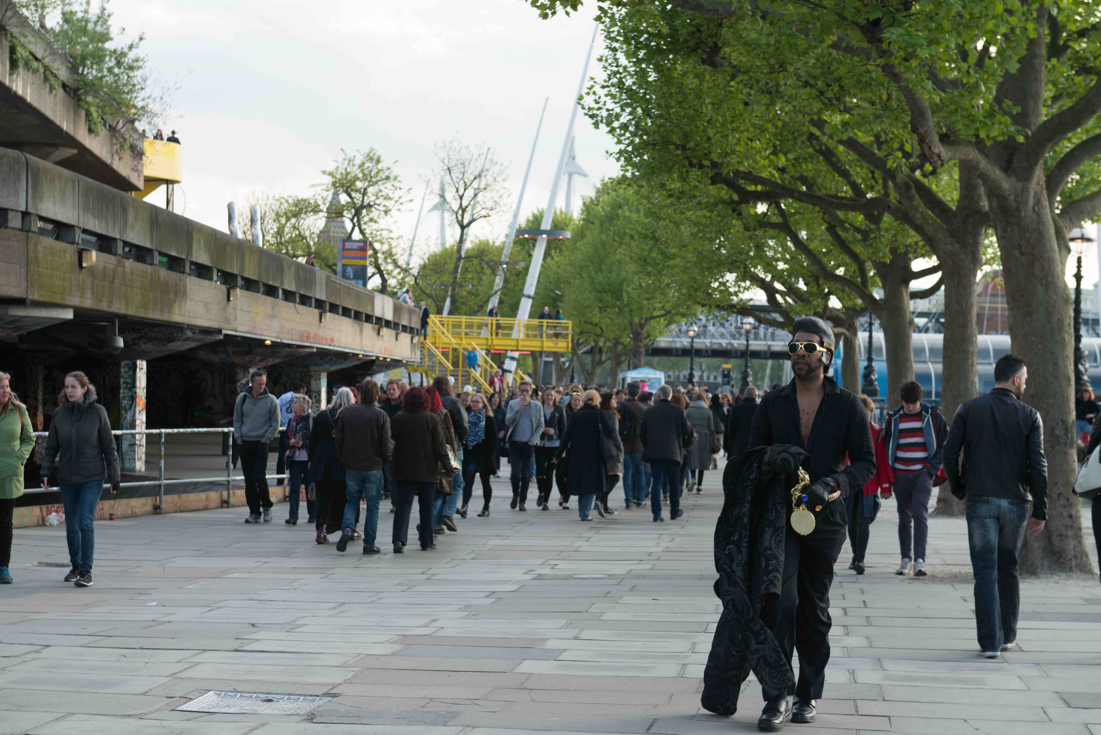Black Elvis impersonator, walking along the southbank with black sunglasses, Black coat and gold medallion and watch. People walking by, street photography london. Instagram.com/vcphoto1
