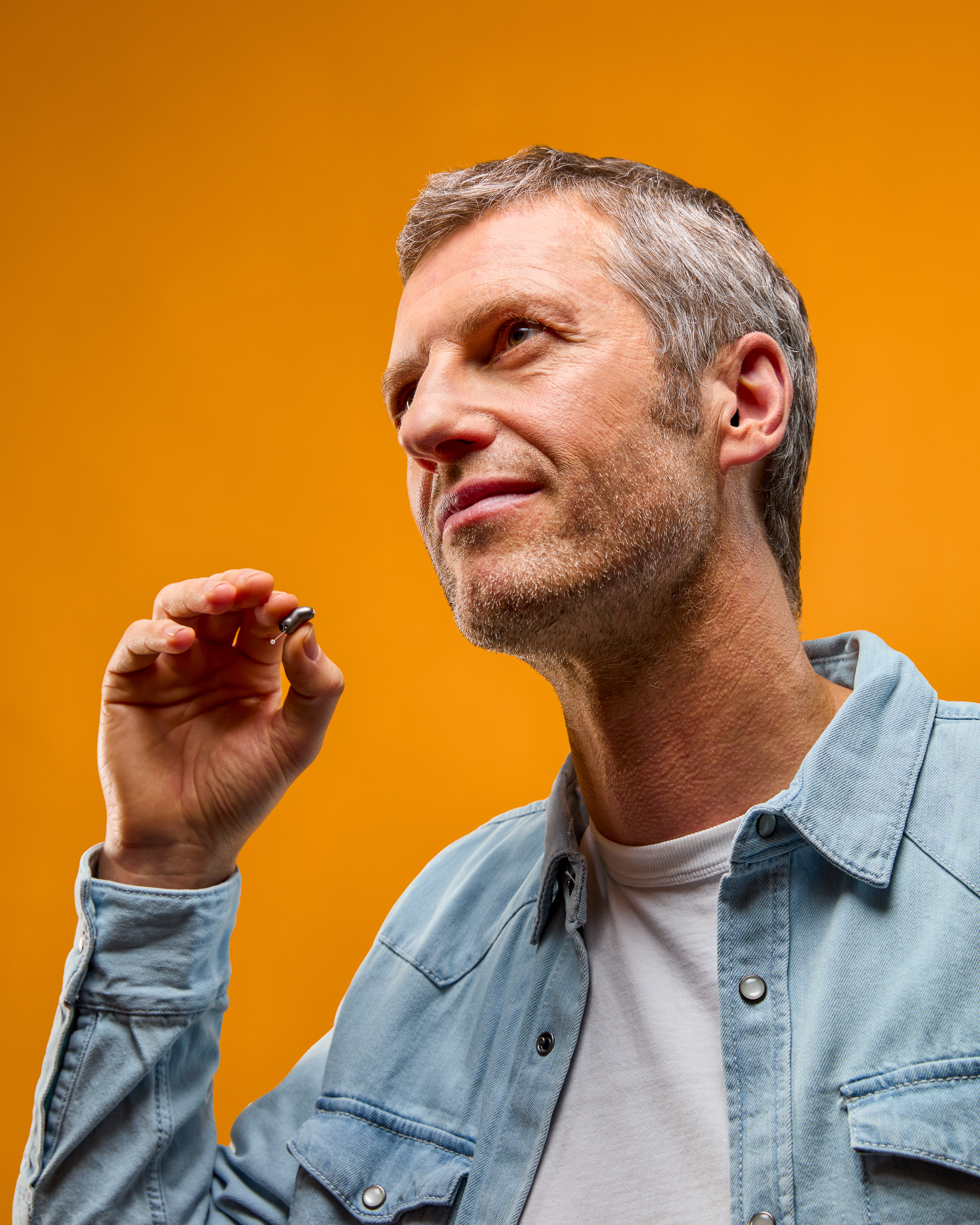 Homme en chemise en jean tenant une aide auditive entre ses doigts, fond orange, studio