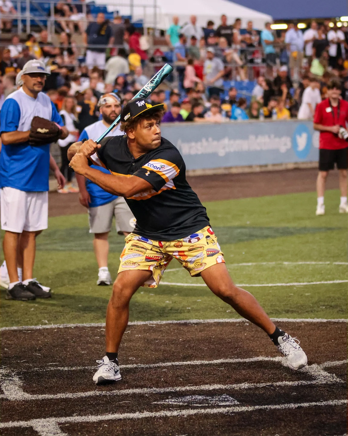 Connor Heyward ready to swing a bat wearing our all-over print shorts, a black jersey, and black and yellow hat.