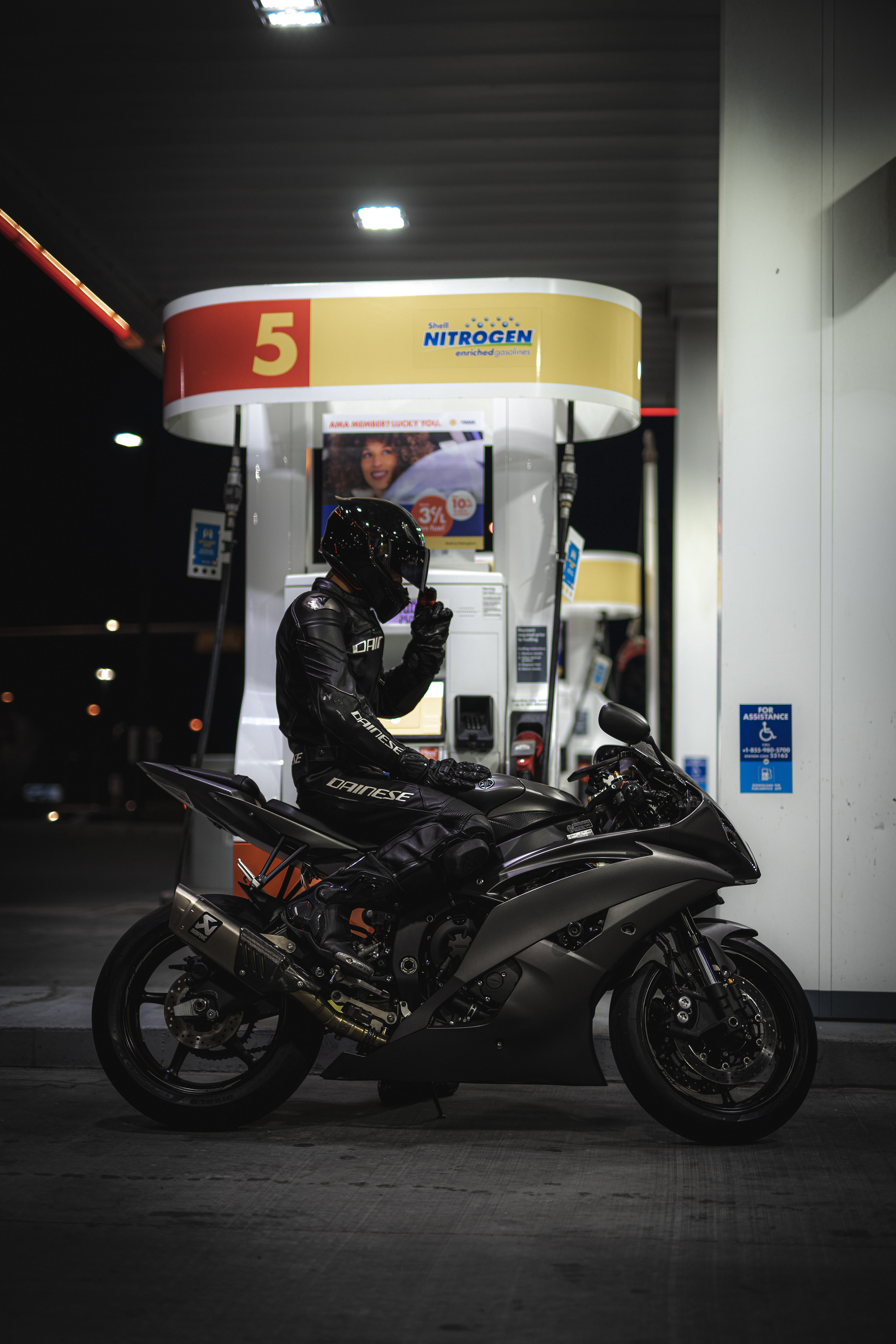 Motorcycle rider in full black Dainese gear sitting on a matte sportbike at a Shell gas station at night, under dramatic lighting.