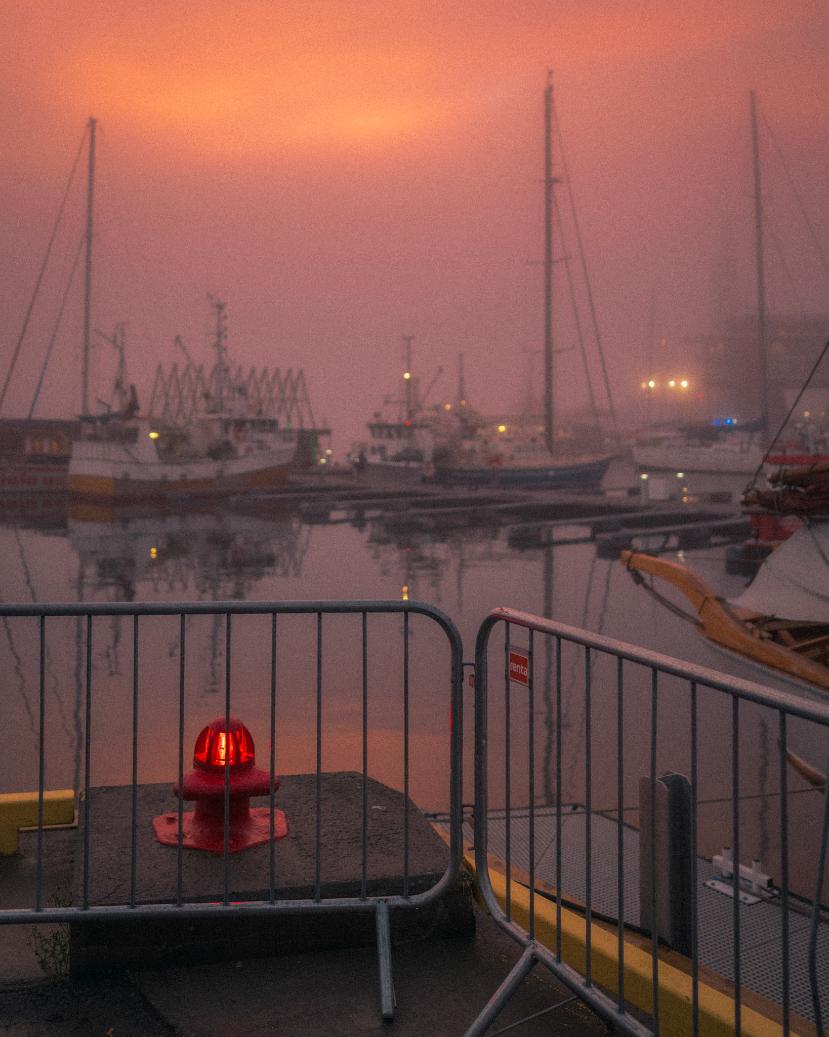 A lone red beacon stands watch over the still harbor, its glow barely cutting through the thick morning fog. The sky burns with the last embers of dawn, casting the boats in a dreamlike haze. A quiet threshold between movement and stillness, warning and welcome.