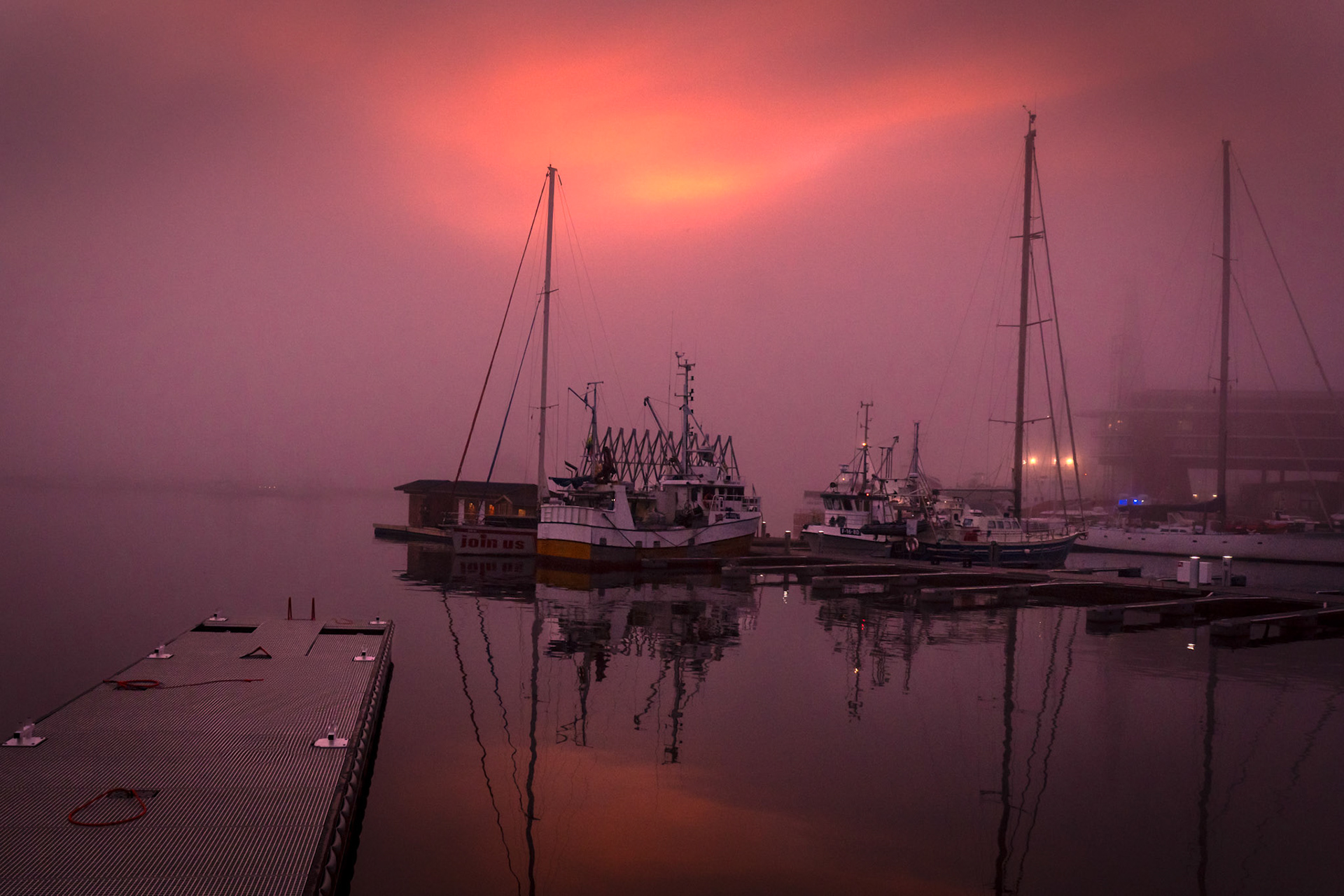 A silent harbor swallowed by fog, where the first light of day rises like a whispered warning. The water mirrors the sky’s muted fire, turning the boats into silhouettes adrift in an ethereal void. This moment, caught between stillness and awakening, feels both cinematic and otherworldly—an invitation into the unknown.