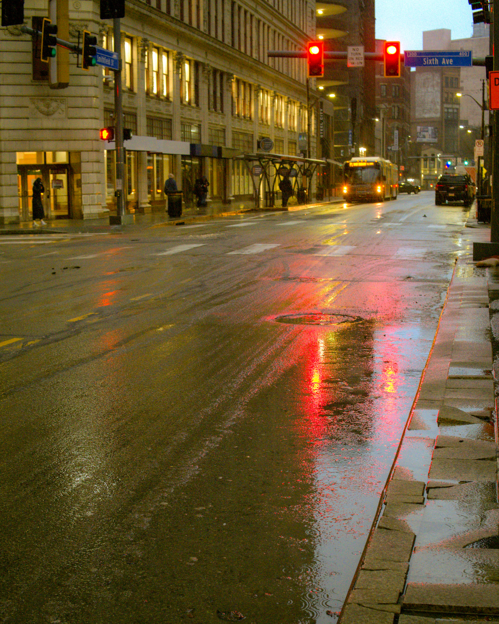 The city pauses in the rain, its rhythm slowed but never stopped. Reflections bleed into the pavement like a painter’s brushstroke, neon reds and golds smearing across the wet asphalt. A bus approaches, headlights flickering in the mist—just another fleeting moment in the endless urban current.