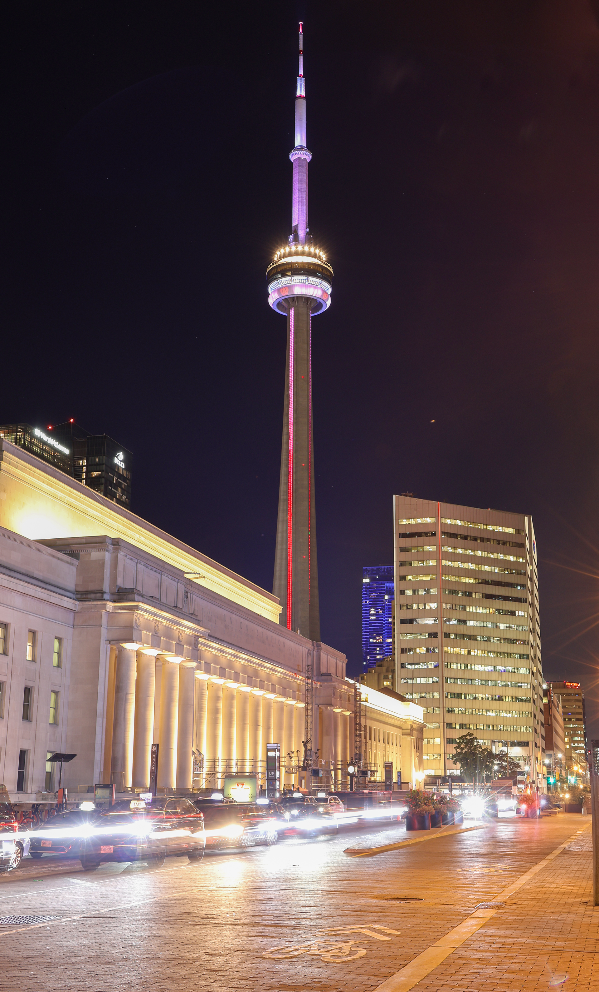 Night view of the CN Tower in downtown Toronto glowing above busy city streets.