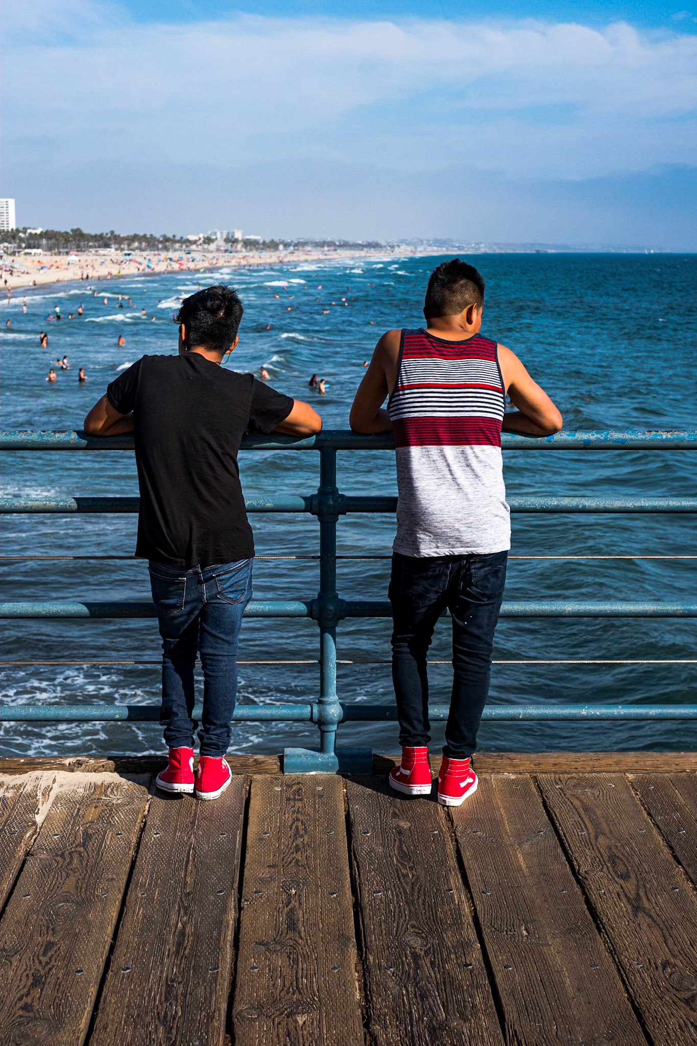 (Mostly) Bicycling America XXXVII. Two Kids Looking Out Over the Pacific at the Santa Monica Pier, Los Angeles, California, August, 2019. I was in Los Angeles after returning from my perambulations abroad because I had left my bicycle with a friend in the city before flying to Australia the previous December. I spent a week wandering around the city getting myself prepared for the bike trip I was planning to take up the coast to the Bay Area. Here I was just out and about photographing. Had never been to the Santa Monica Pier before, and thought it a good idea to visit while I was in town. With their red shoes and similarly relaxed leaning on the railing, looking in opposite directions over the water, this was too good a picture to pass up, even if it is slightly off-center.