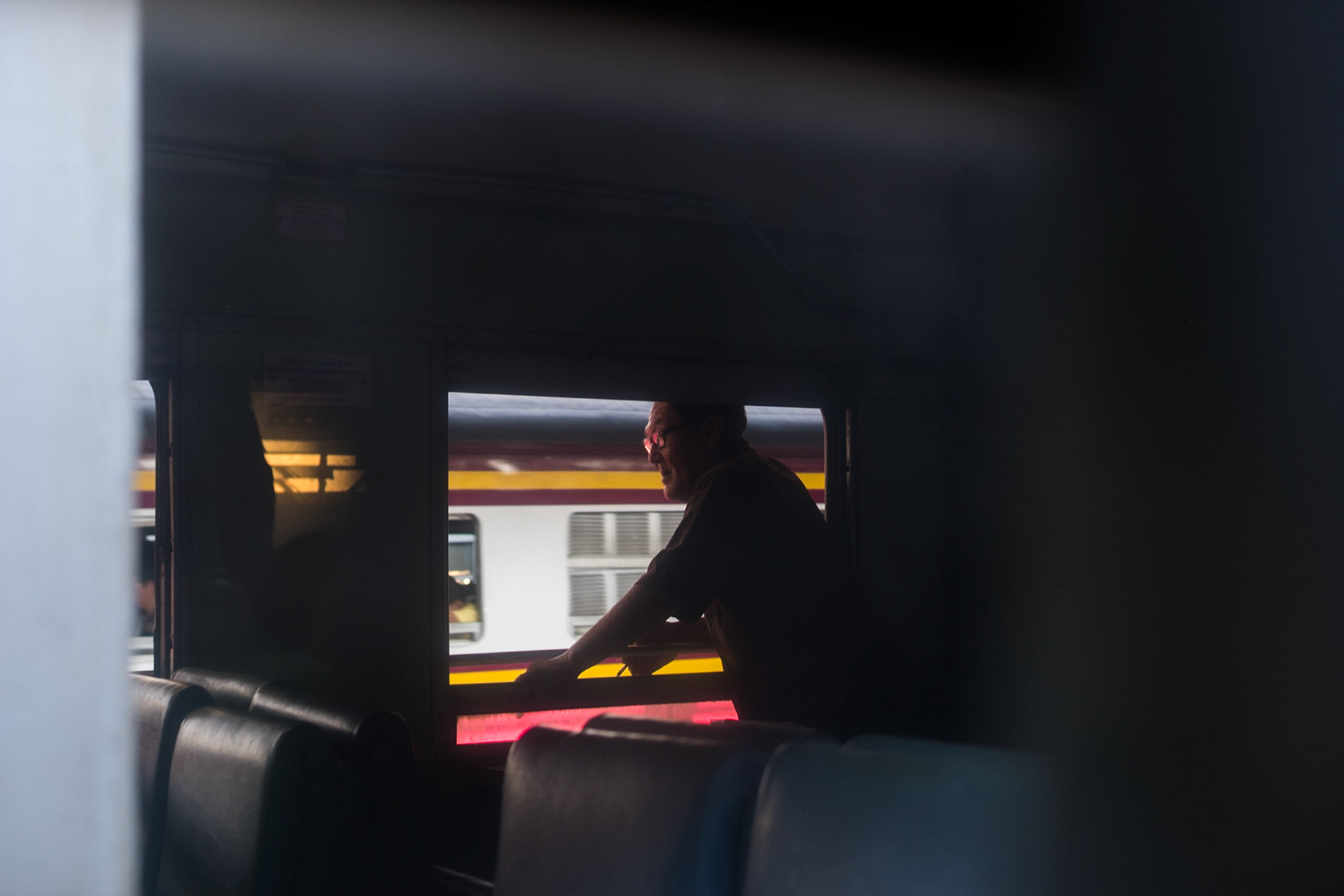 The Rapture of Train Travel II. A man smokes a cigarette while leaning through a window of a train before departure at Bangkok train station.