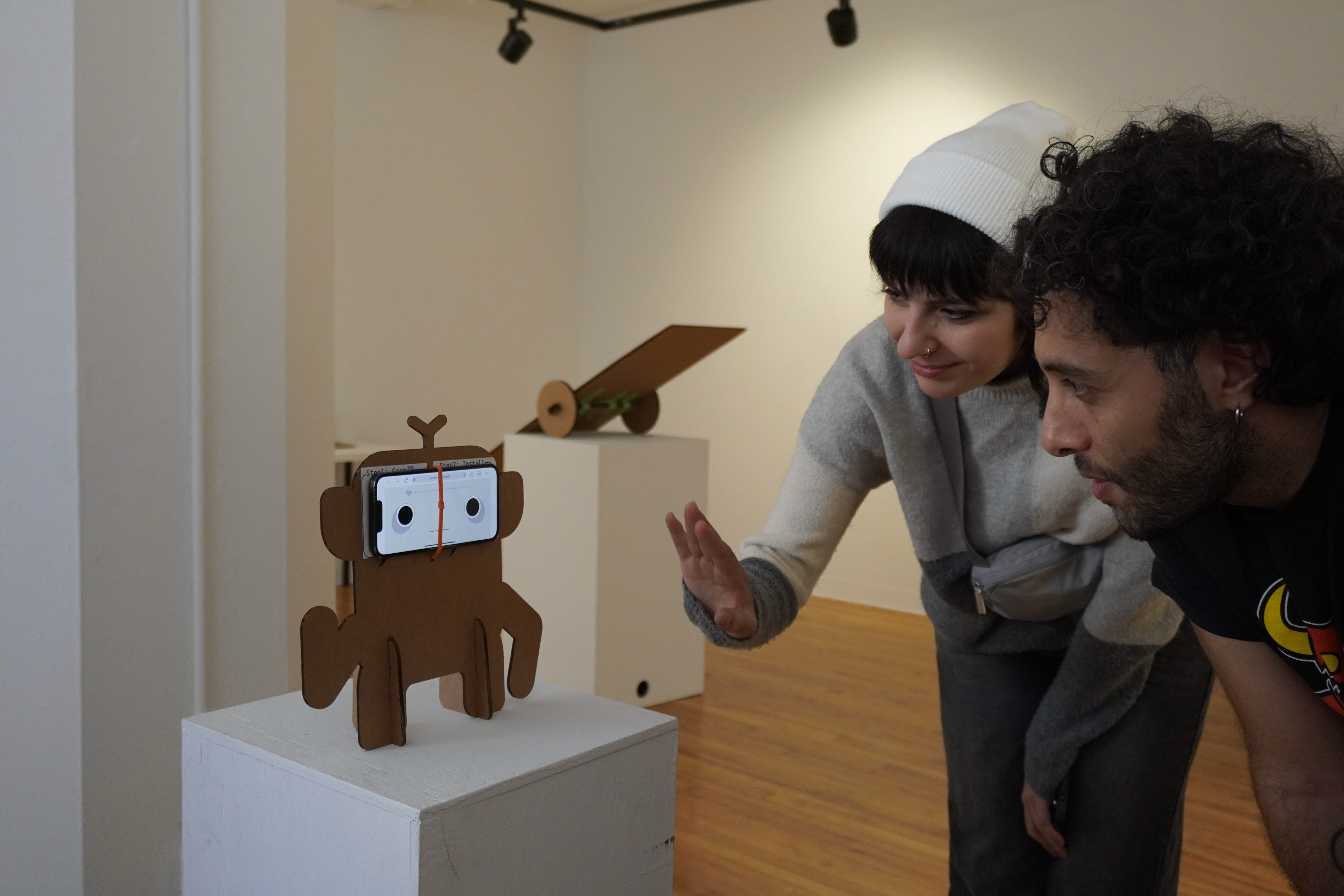 Image of phone pet on a plinth with two people interacting with it. Several people placed their phone on a plinth for others to come up and interact with it, which was really fun to see humans attempt to emulate the pitches to communicate with the pet.