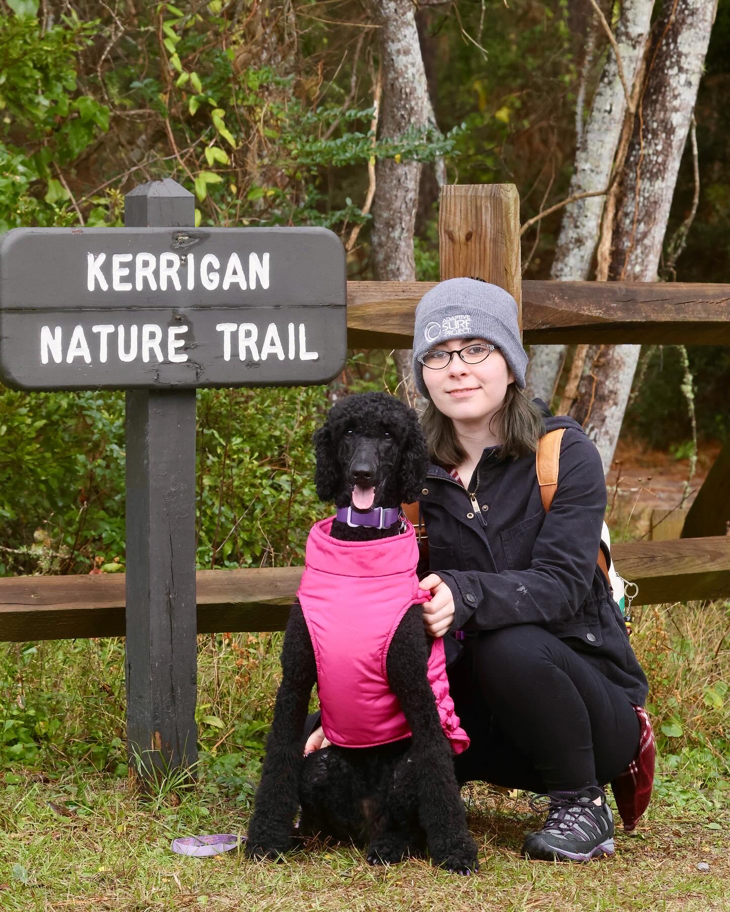 Eleanore, a white woman, is wearing a winter hat, a light coat, and a backpack. She is crouching next to her black standard poodle, Jinkx, who is wearing a pink winter vest. They are posing next to a sign that says Kerrigan Nature Trail, and there is a wooden fence, trees, and other foliage in the background.