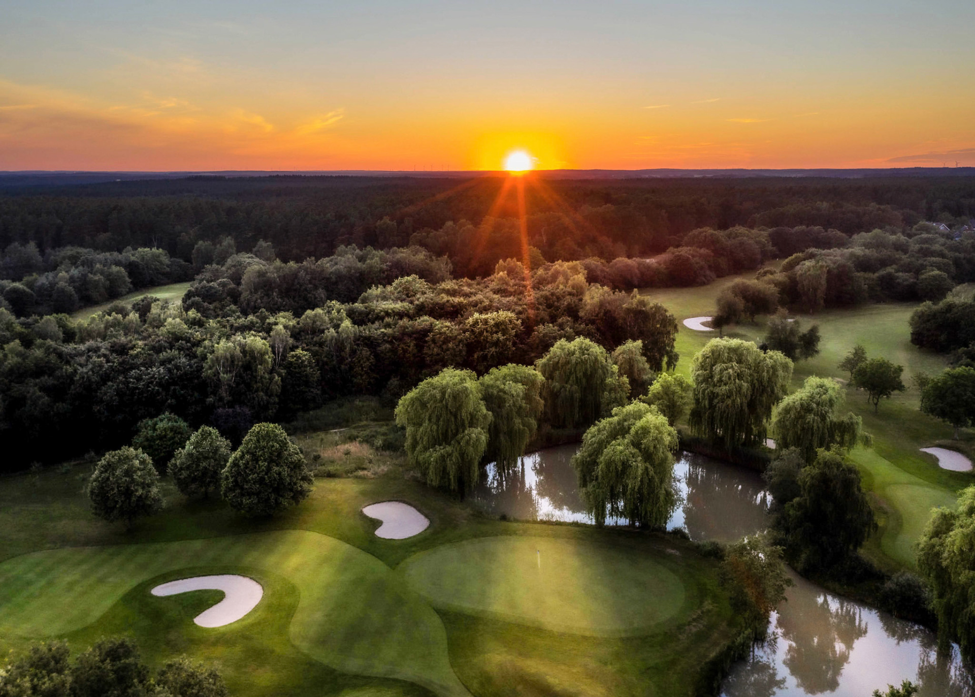 Atemberaubender Sonnenuntergang über dem 18-Loch Parkland Golfcourse Bad Bevensen. Sonnenstahlen dringen durch den alten Baumbestand und erleuchten das GRün der Bahn 9. Einem Par 3 direkt vor dem Clubterrasse des Clubrestaurants HELD's. Der ideale Ort, um seine Golf-Platzreife zu machen.