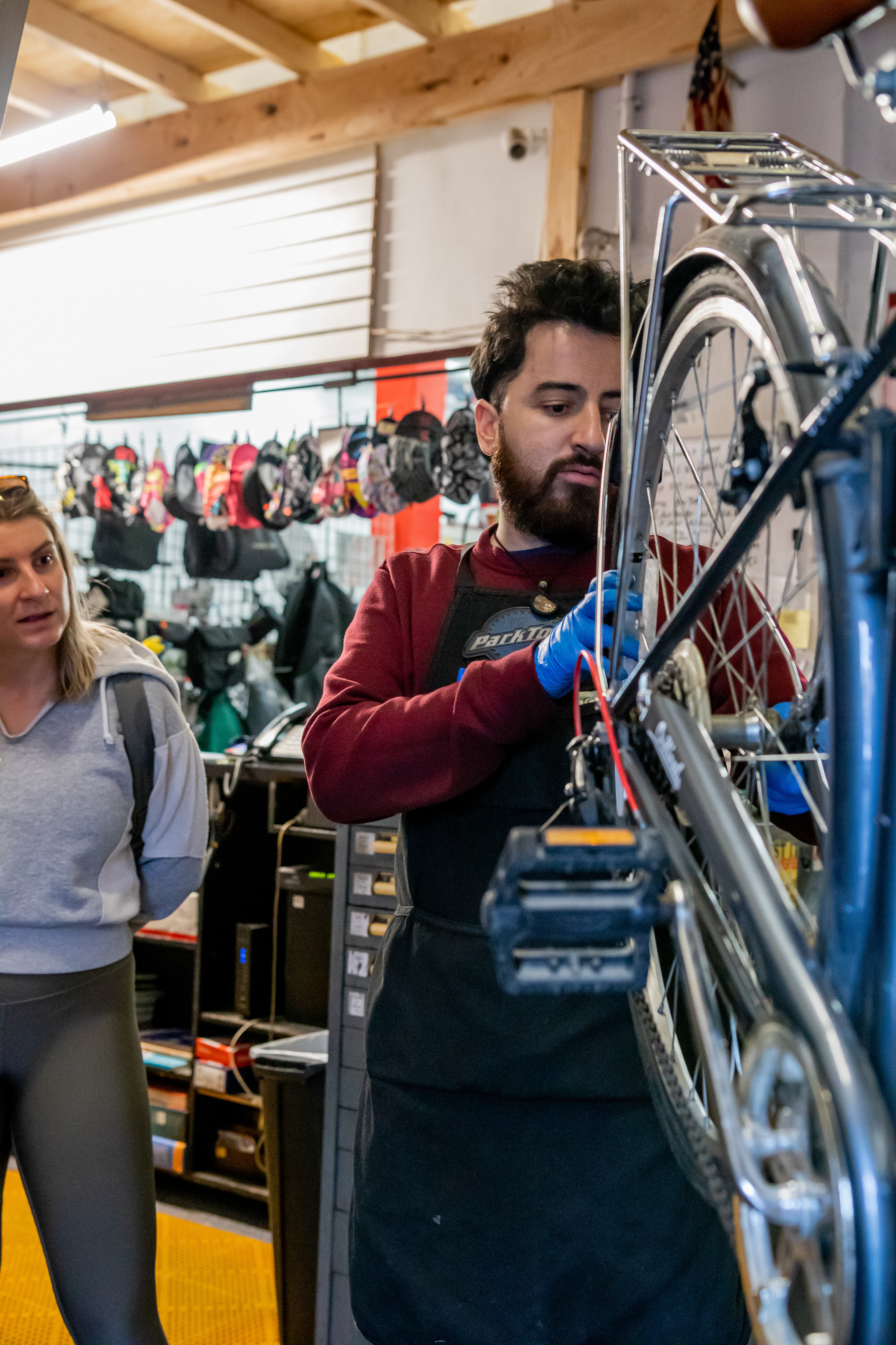 A customer speaks to mechanic Fabian Gil regarding a repair as Gil inspects her bike, April 15, 2022 at Community Bike Supply, Boston, Massachusetts. As the weather warms, business starts to pick up for the mechanics as people bring their bikes out for tunes and repairs before the summer season (Ryan Li).