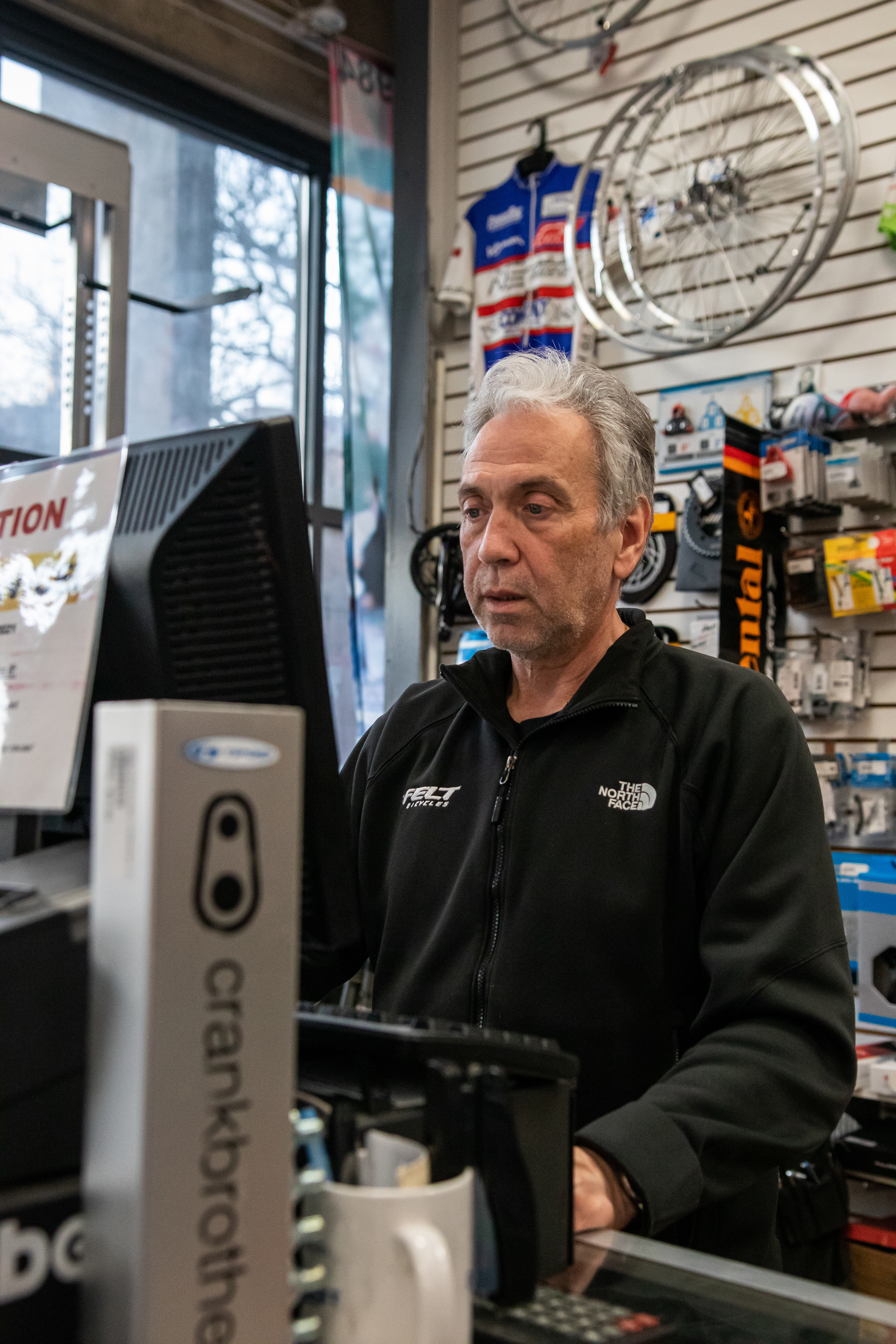 Phil Coombs stands behind the front desk checking orders for inventory, April 15, 2022 at Community Bike Supply, Boston, Massachusetts. Phil mainly works in the front, ringing up customers and helping whomever passes through the store. He has worked at Community for 42 years, but is nulling retirement soon to join his wife in Florida (Ryan Li).