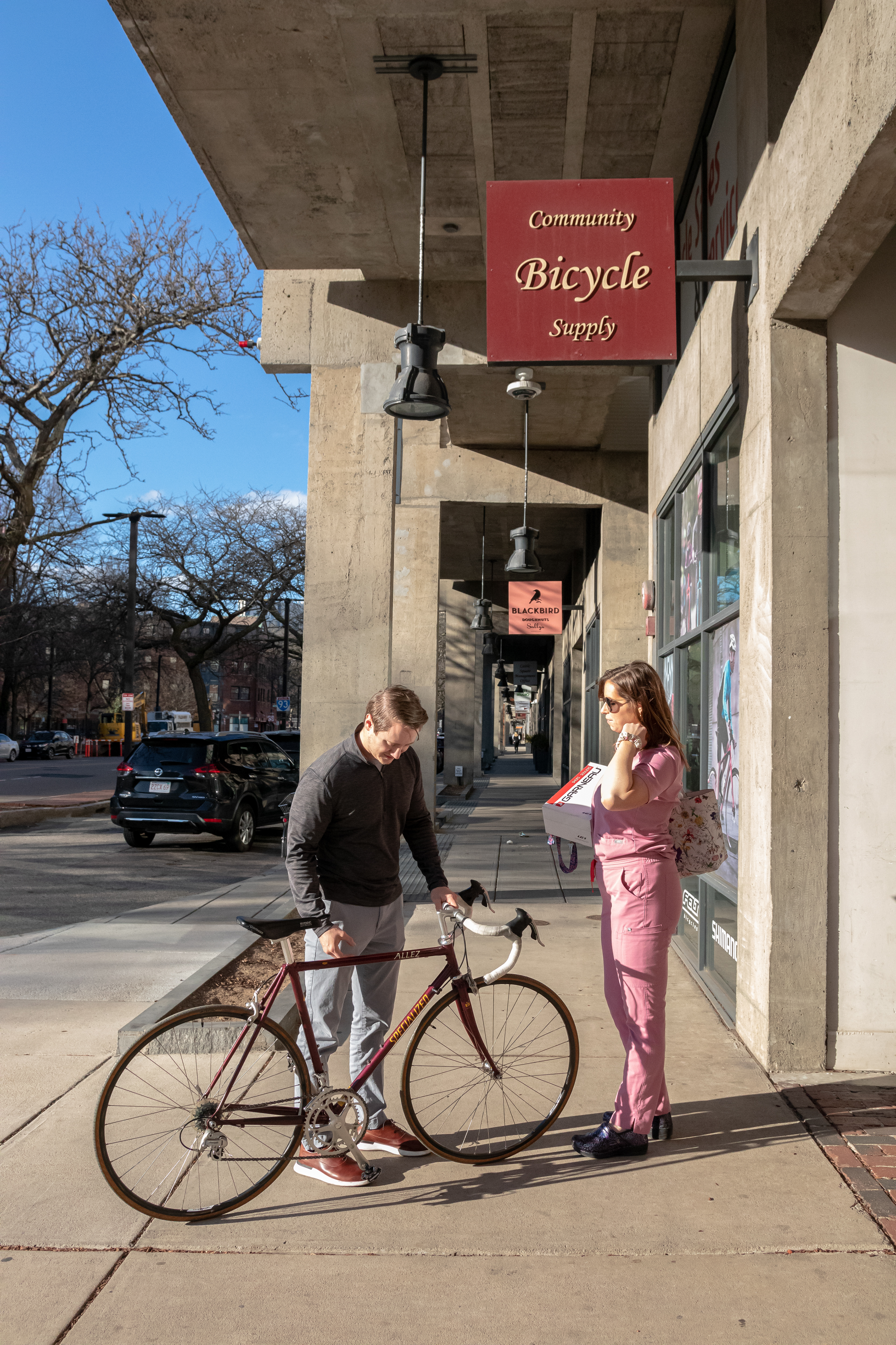 A couple stands outside of the shop doors after picking up a repair and purchasing a pair of biking shoes, April 22, 2022 at Community Bike Supply, Boston, Massachusetts. They mentioned that they had purchased the bike the day before off of Facebook Marketplace and brought it in to get cleaned and tuned up for an upcoming bike race (Ryan Li)