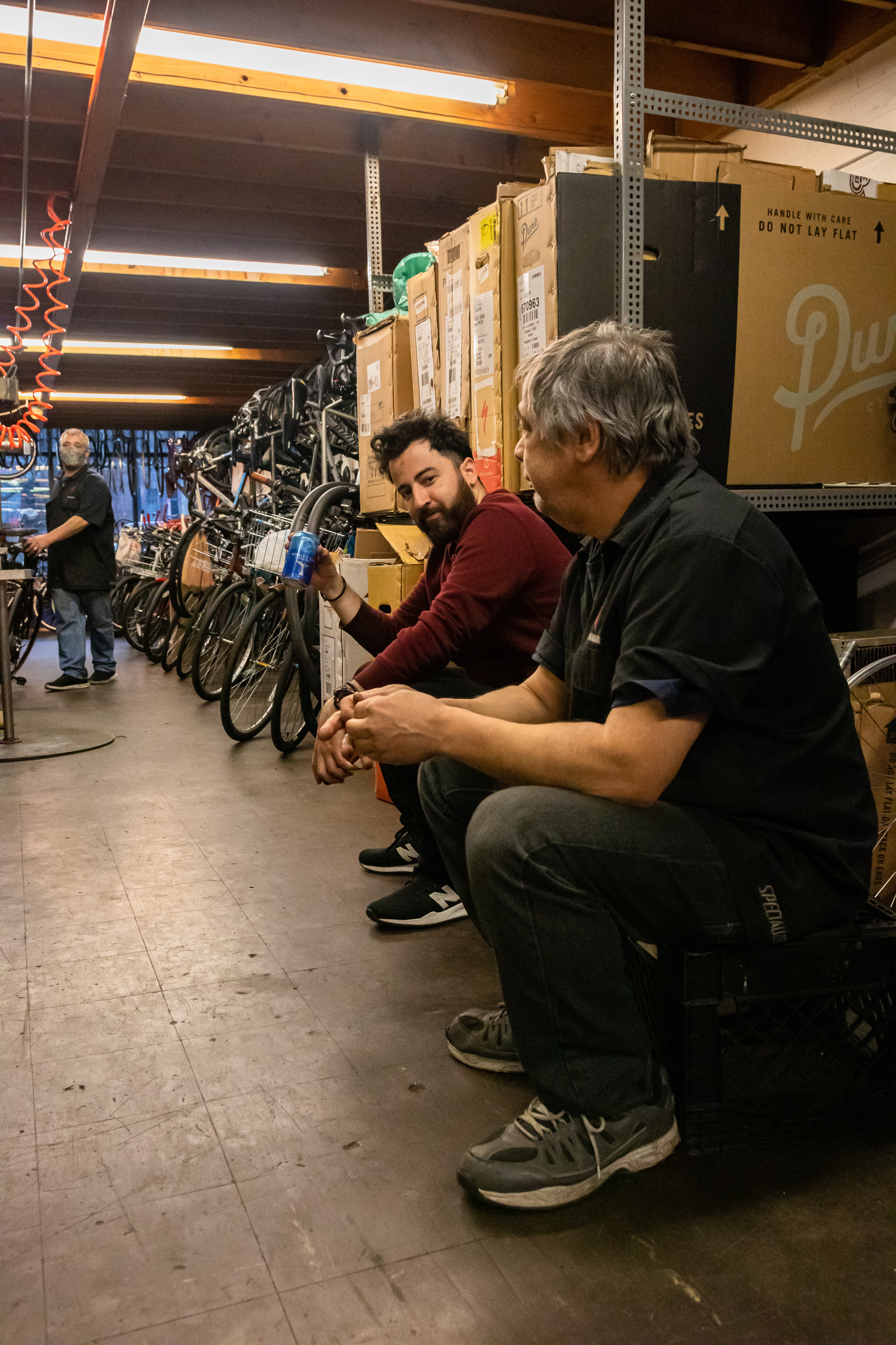 Fabian Gil and the other bike mechanics sit on crates in the warehouse after store closing to enjoy a beer and socialize, April 15, 2022 at Community Bike Supply, Boston, Massachusetts. The mechanics are a tight knit group and enjoy poking light fun at each other (Ryan Li).