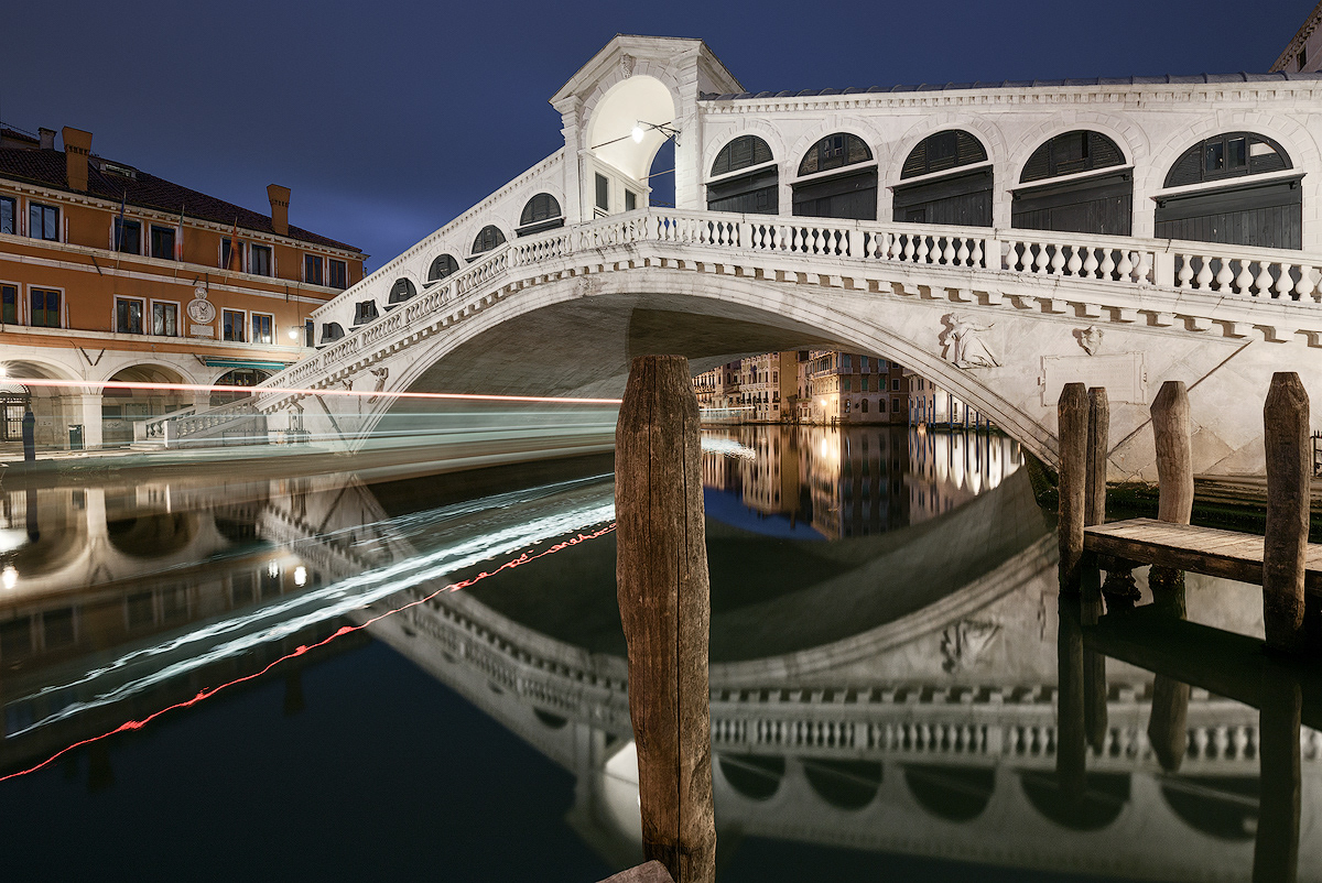 Ponte di Rialto a Venezia fotografato di notte in lunga esposizione, con scie luminose di un vaporetto sul Canal Grande e riflessi sull'acqua