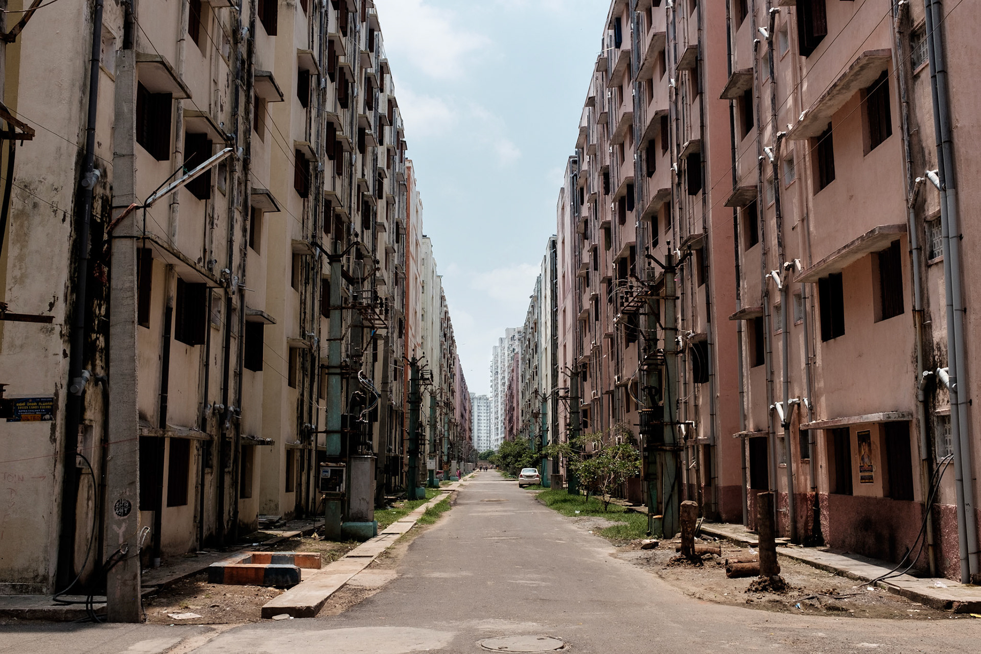 These housing blocks are being built on the fringes of Chennai city. While the hosuing might look spare and isolated, it is nonetheless an improvement over living on a pavement or in a slum.
Within these blocks are small shops, skills training centres, and community actrivities. The housing blocks are being built next to more expensive housing for office and professional workers, creating an economic ecosystem that will provide jobs such as domestic workers, drivers, and other services to their wealthier neighbours.
The white buildings at the end of this street are middle-income housing blocks.
