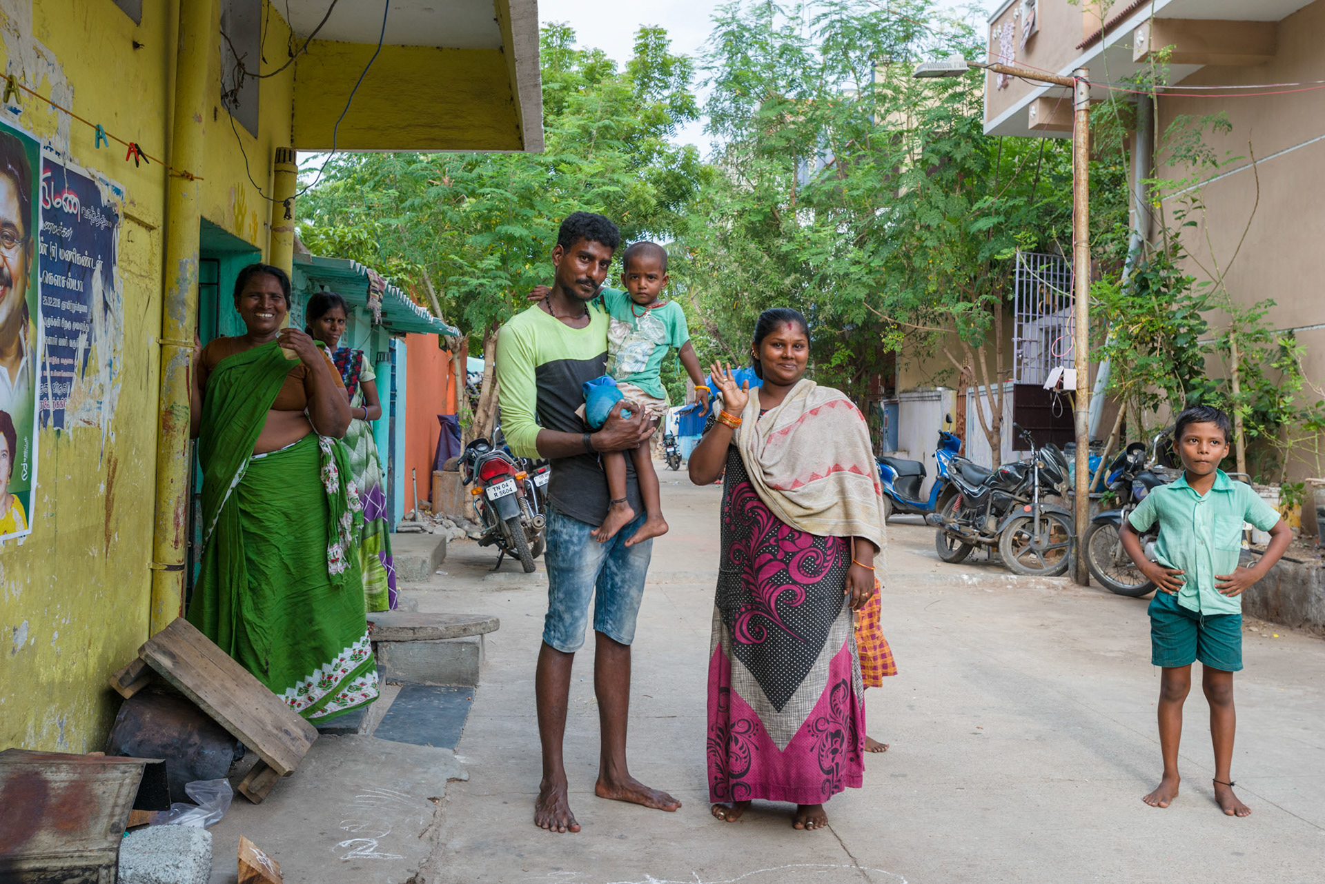 Sayagamari, with her husban Kuppuswamy, and daughter wave ouy side their apartment.
A little over a year ago, Sayagamari lived on the pavement with her husband and duaghter. Since then she has moved to permanent accomodation. She still works with her husband selling drinking water, but now she has a secure place to live on a good street. She is safer, healthier, and happier.g