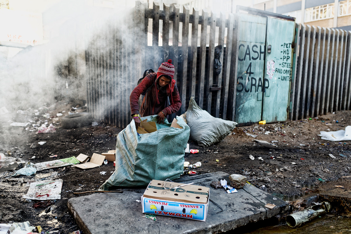 Recycling Outside Dark City 2014 - 2016 Photographic Print Fine Art Cotton Rag A1. A man recycles boxes on a street corner outside of Dark City. The fire in the background is from burning of cables to sell copper for scrap metal.
