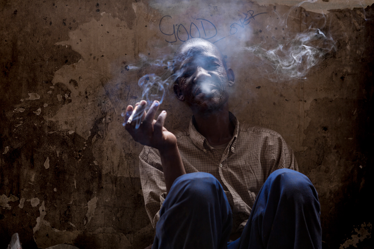 James, Low Portrait 2014 - 2016 Photographic Print Fine Art Cotton Rag A1. Portrait of James an immigrant from Zimbabwe resting against a wall smoking a cigarette in Dark City.