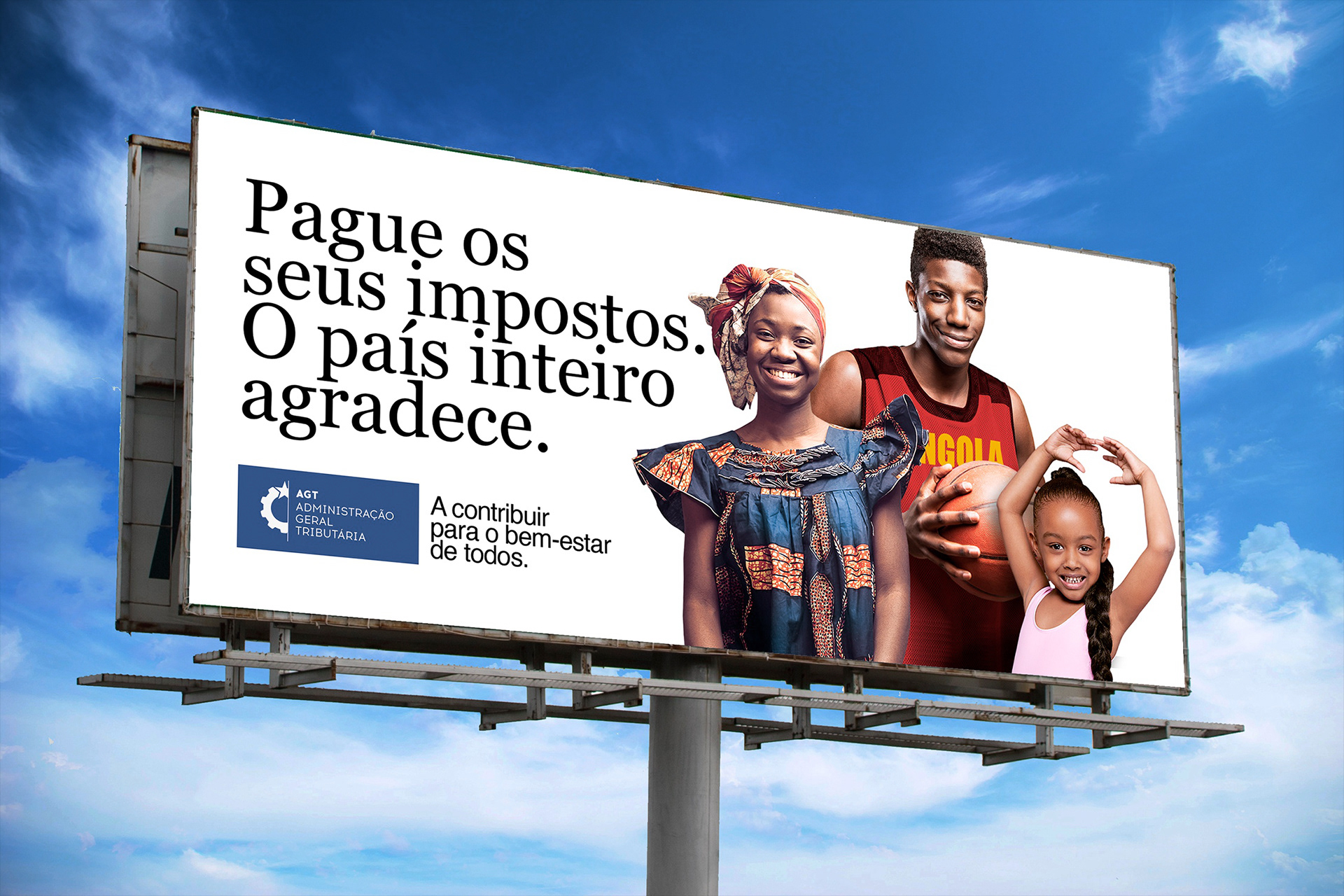 A billboard with a blue sky background displays a message in Portuguese urging people to pay their taxes for the nation's benefit. It features the AGT Administração Geral Tributária logo and three individuals, each representing different demographics.
