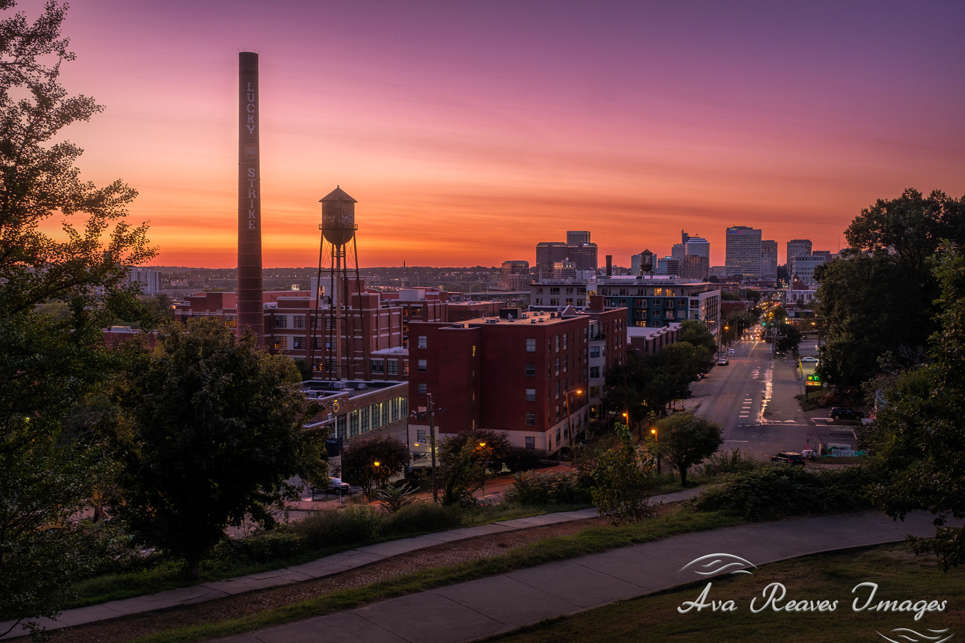 A vibrant sunset view from Libby Hill Park in Richmond, Virginia, featuring the historic Lucky Strike smokestack, water tower, and the city skyline under a colorful purple and orange twilight sky.