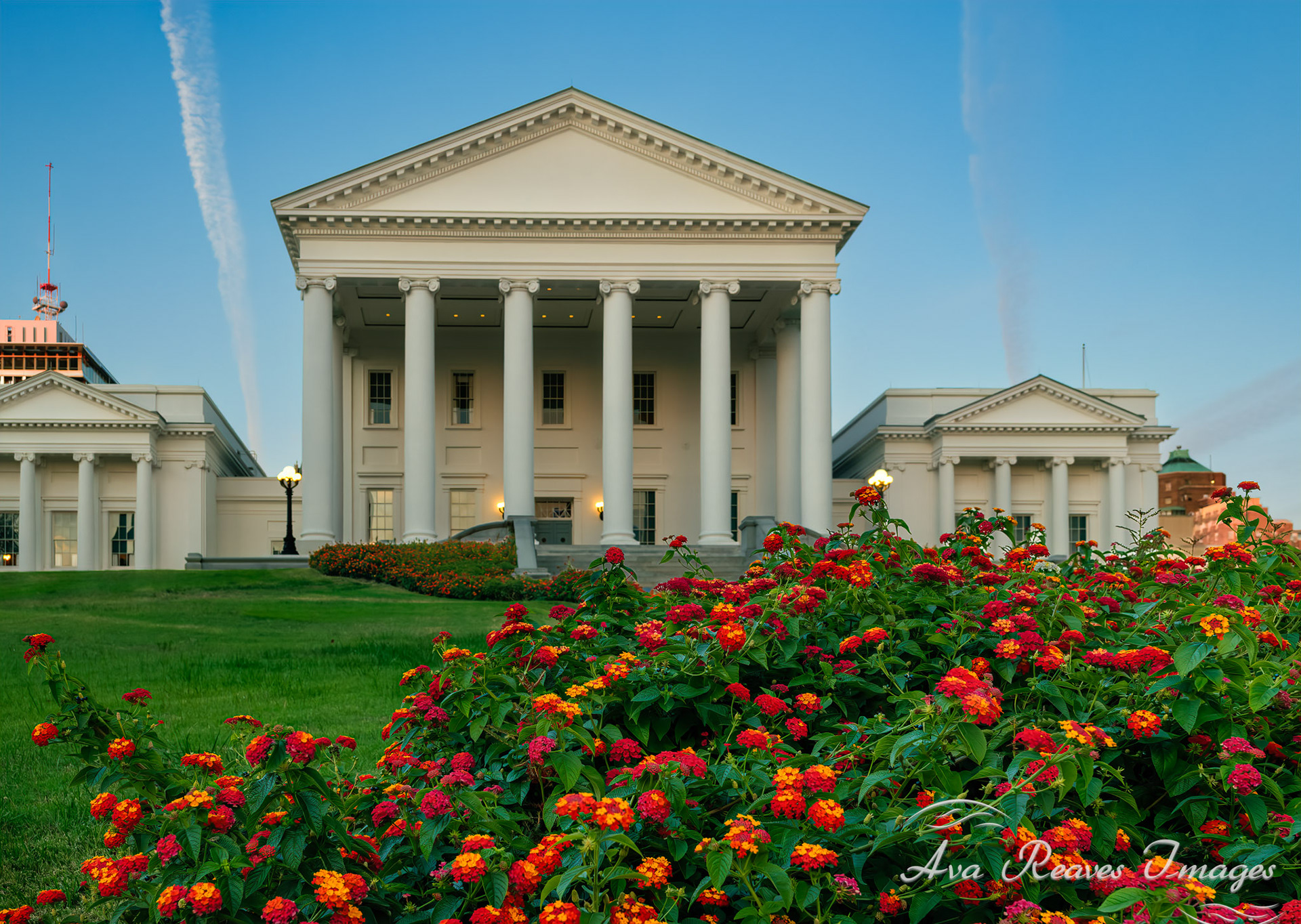 The white neoclassical Virginia State Capitol building framed by vibrant red and orange flowers at dusk