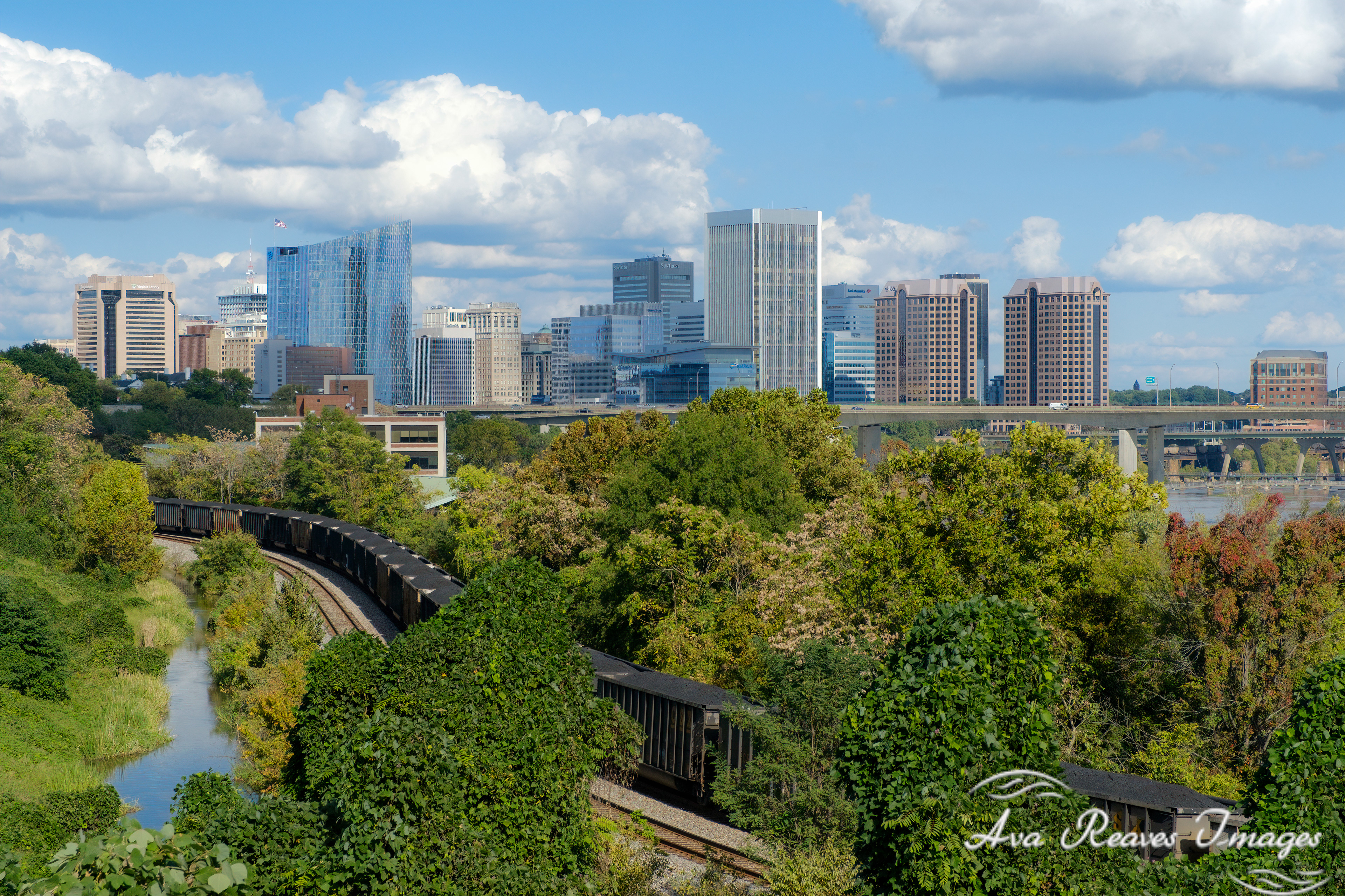 A CSX coal train with multiple black rail cars curves along a track next to the kanawha canal, with lush green trees and foliage in the foreground. In the background, the downtown Richmond, Virginia skyline is visible under a partly cloudy blue sky, with the Lee  bridge spanning the James River.
